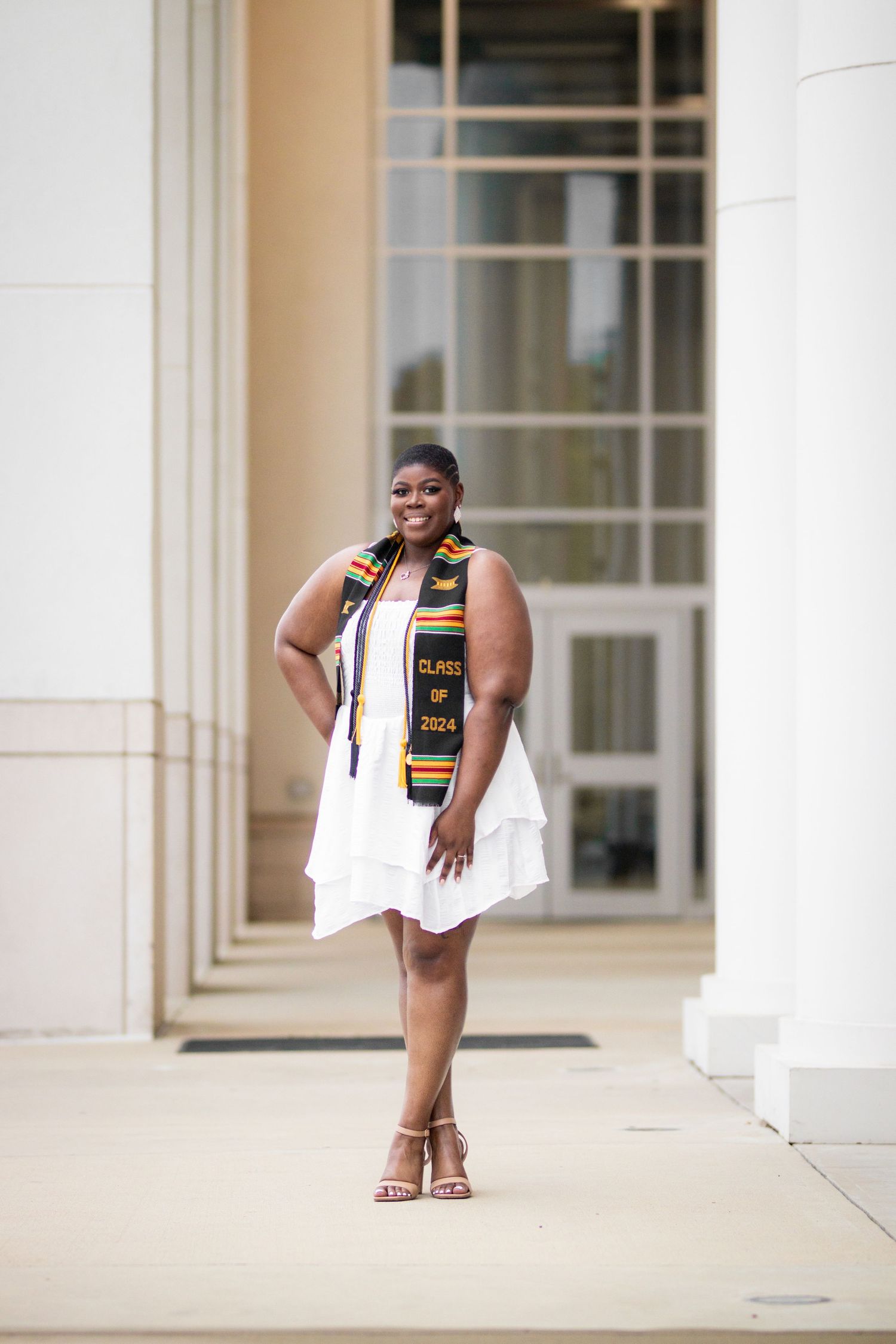 Smiling graduate in white dress and Class of 2024 kente stole poses between columns outdoors.