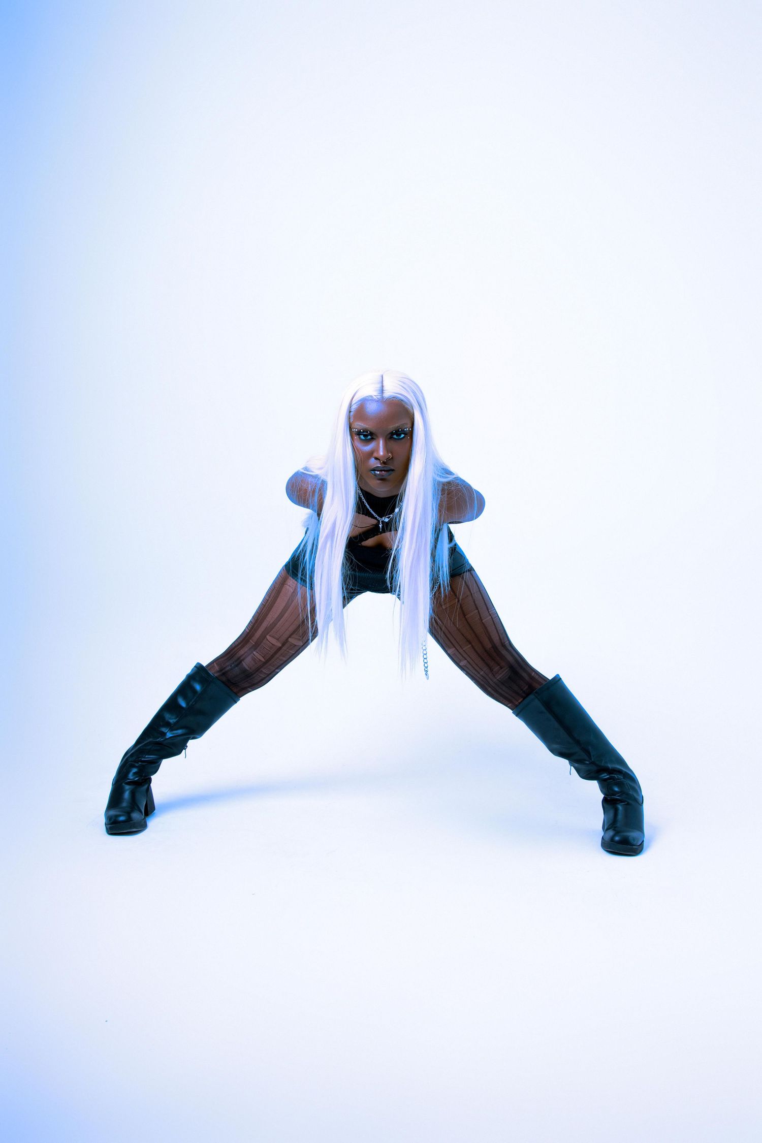 A woman in a black bodysuit and knee-high boots poses in a wide stance with long white hair against a blue-white background.