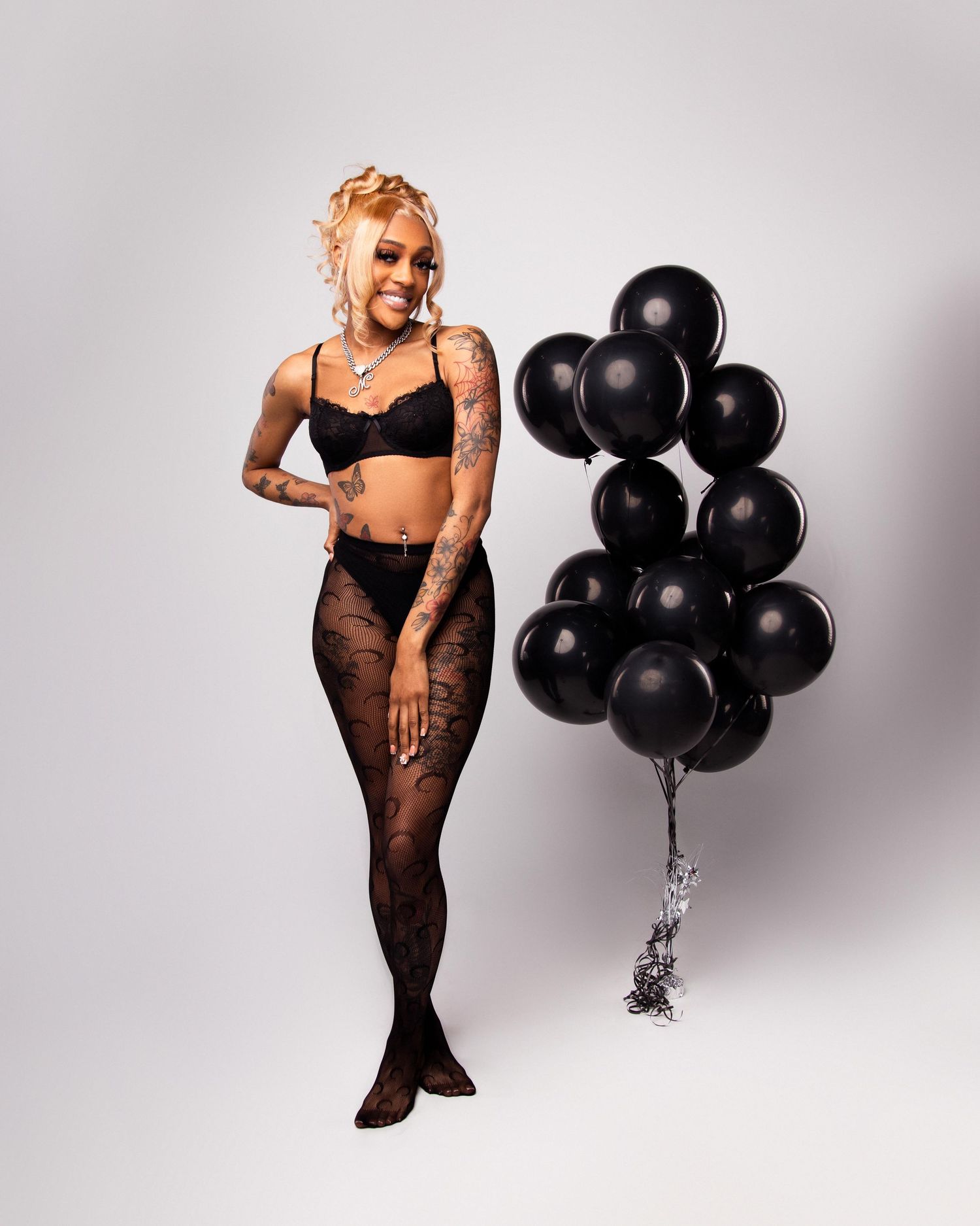 Tattooed woman in black lingerie and tights smiling while holding black balloons against white background.