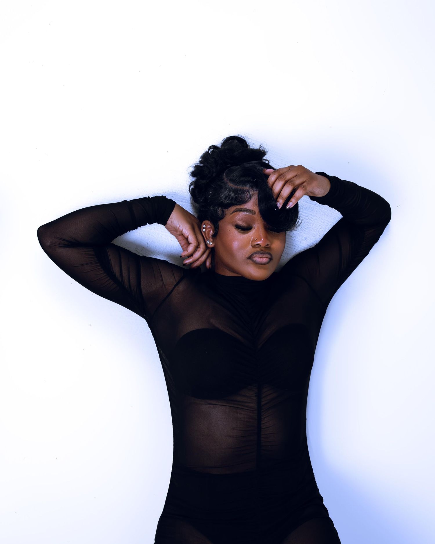 Woman in black sheer long-sleeve bodysuit posing with hands in hair against white background.