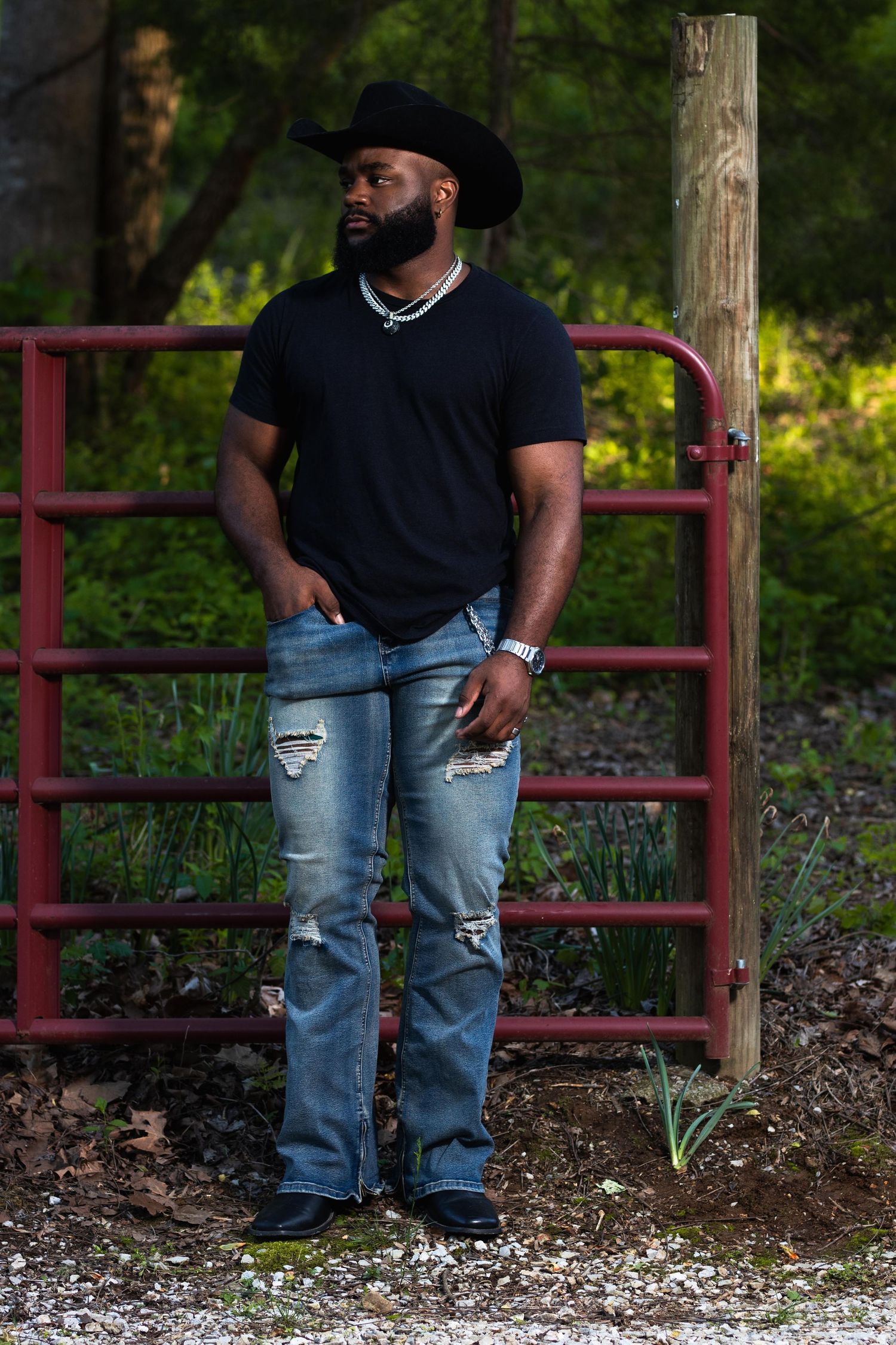 Muscular Black man in black cowboy hat, black t-shirt, and ripped jeans leaning on a red gate outdoors.