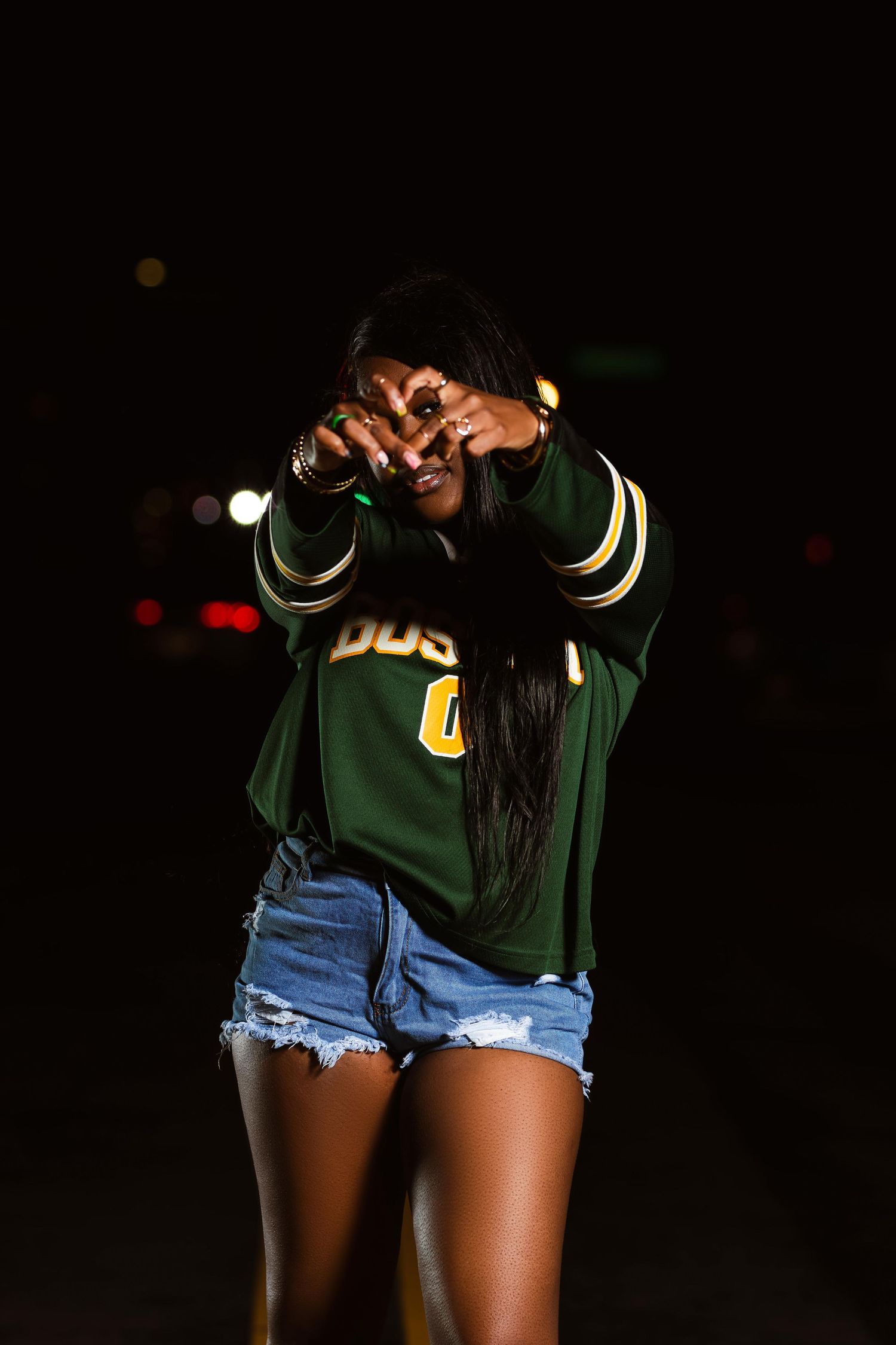 Young Black woman in green varsity jacket and denim shorts making a heart shape with her hands at night outdoors.
