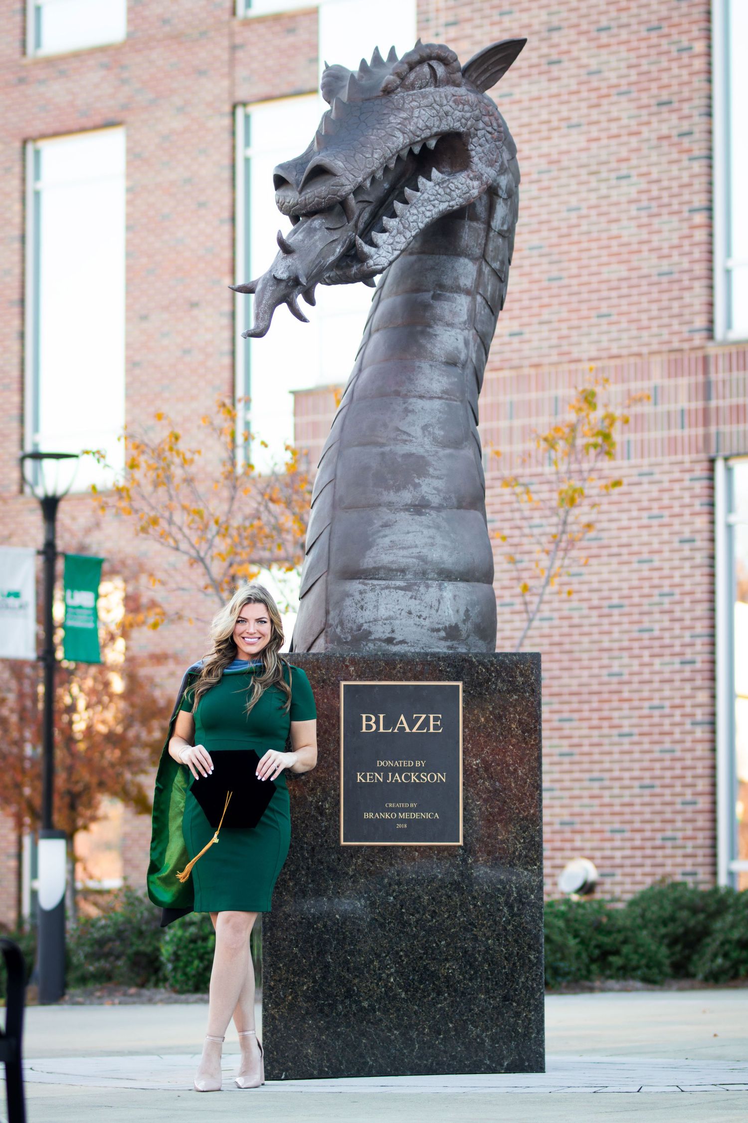 Female graduate in green dress poses beside Blaze dragon statue at UAB.