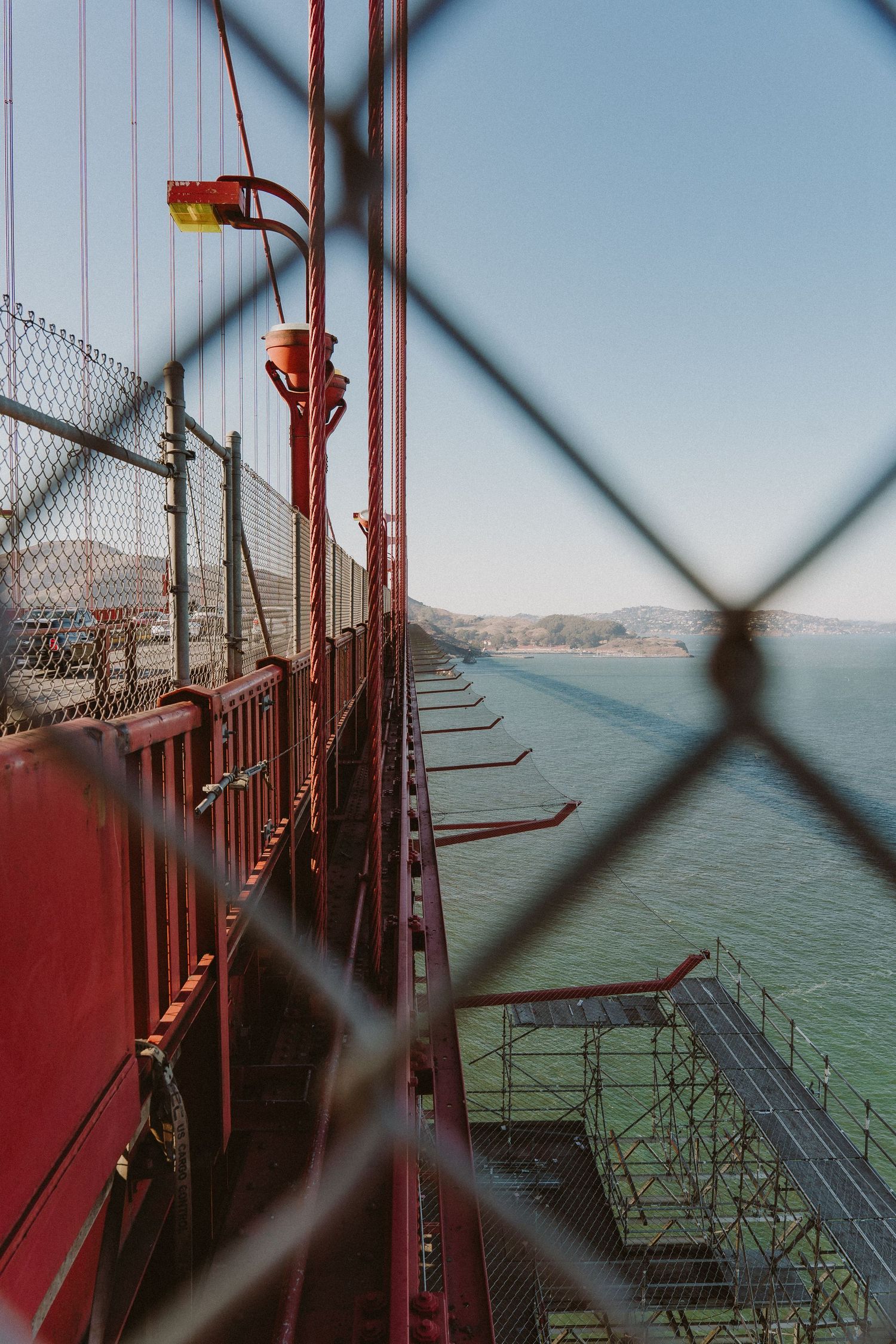 View through safety fence of the Golden Gate Bridge's walkway and support structures on a sunny day, with bay visible below.