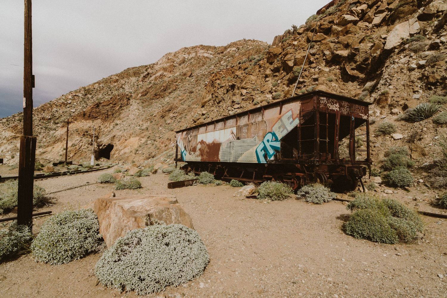 Desert landscape with rocky hills and abandoned buildings against an overcast sky.