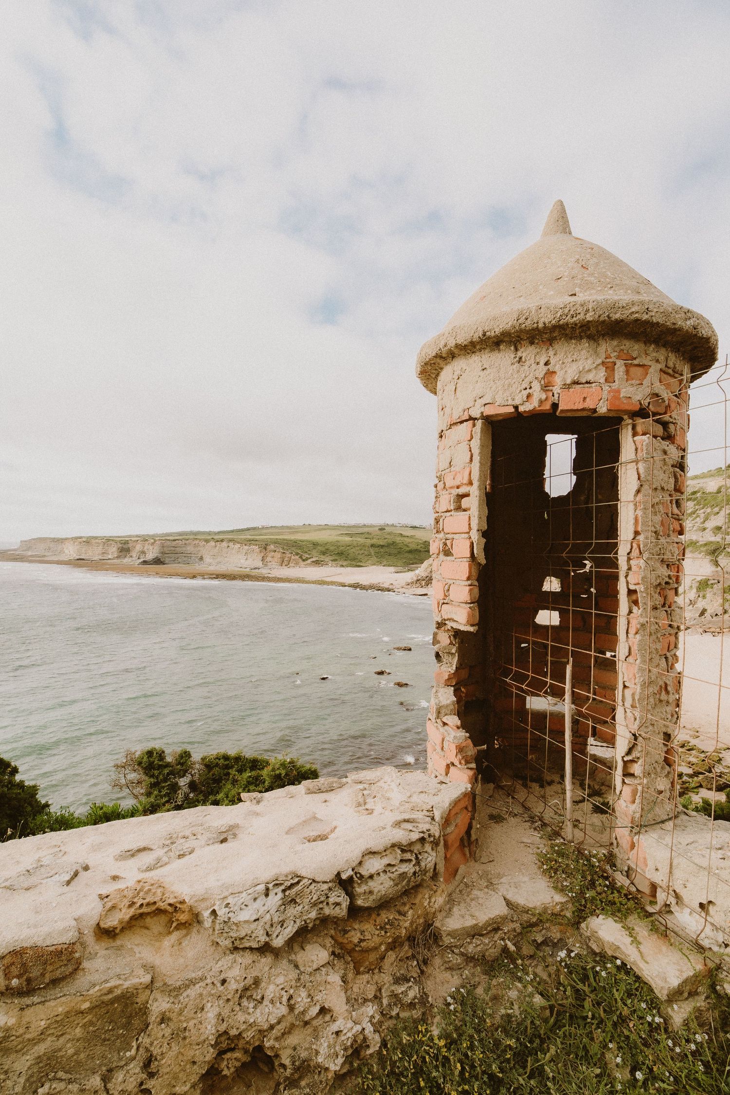 Old stone watchtower turrets overlooking coastal cliffs with ocean views and rolling green hills in the distance.