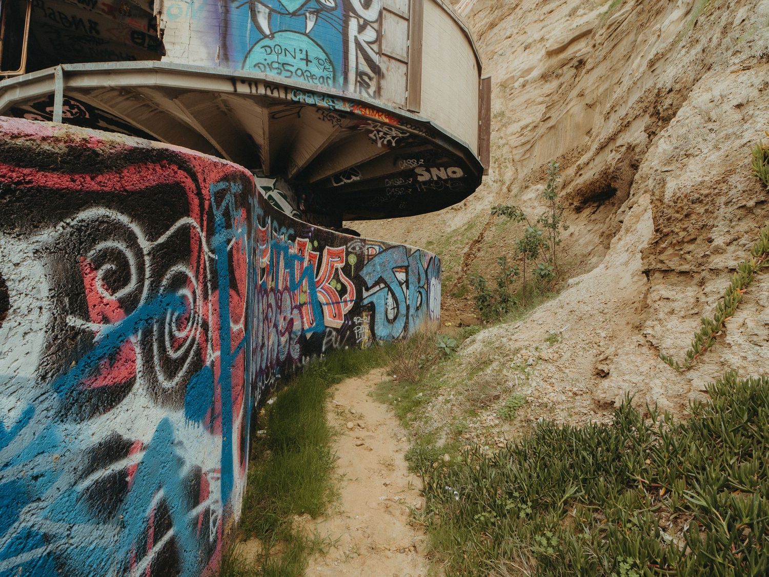 Colorful graffiti art decorates a curved concrete tunnel wall along a dirt path with rocky cliffs in the background.