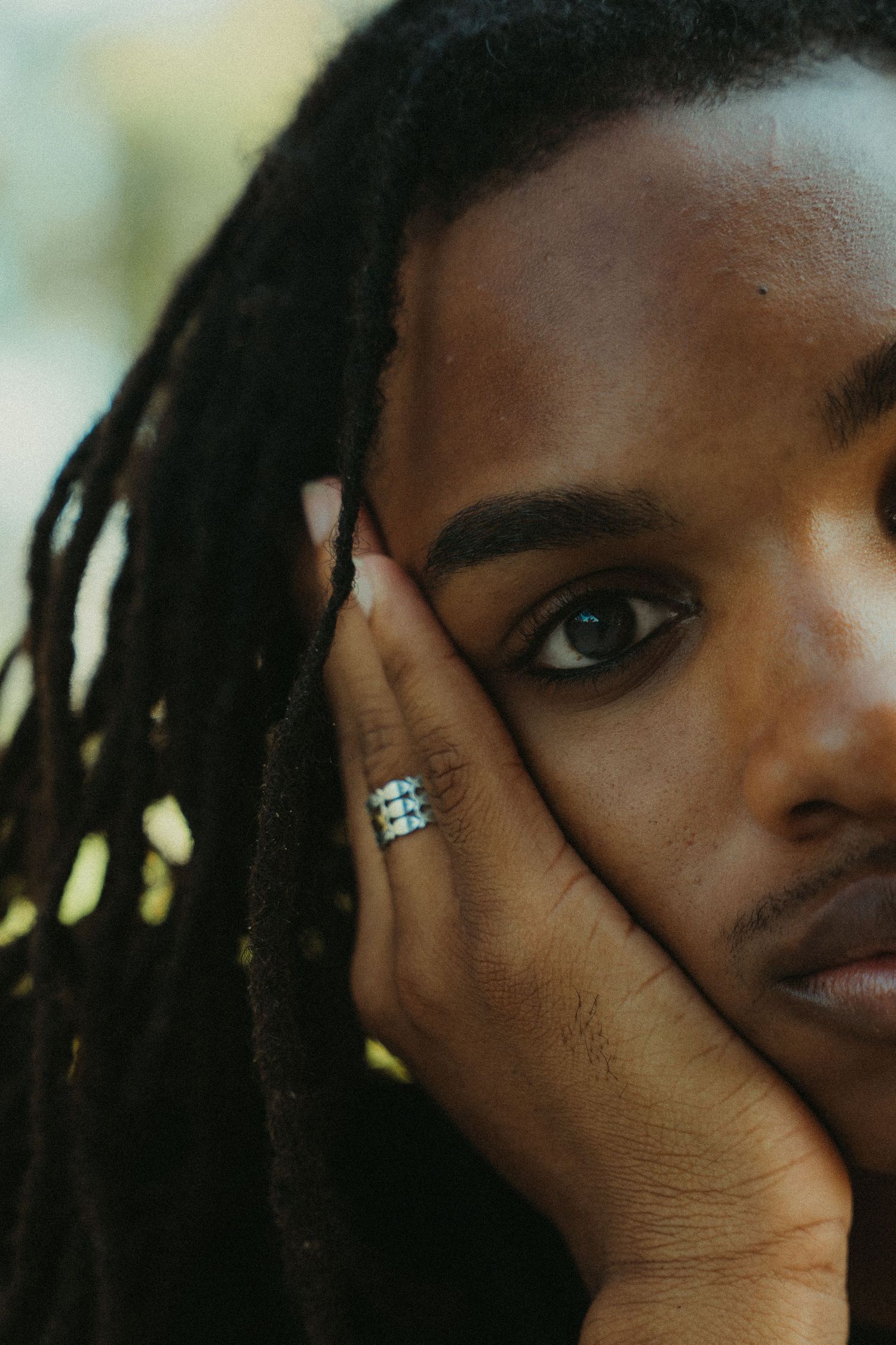 Close-up artistic portrait model with dreadlocks showing one eye and a hand wearing a sparkly ring against a blurred background photoshoot