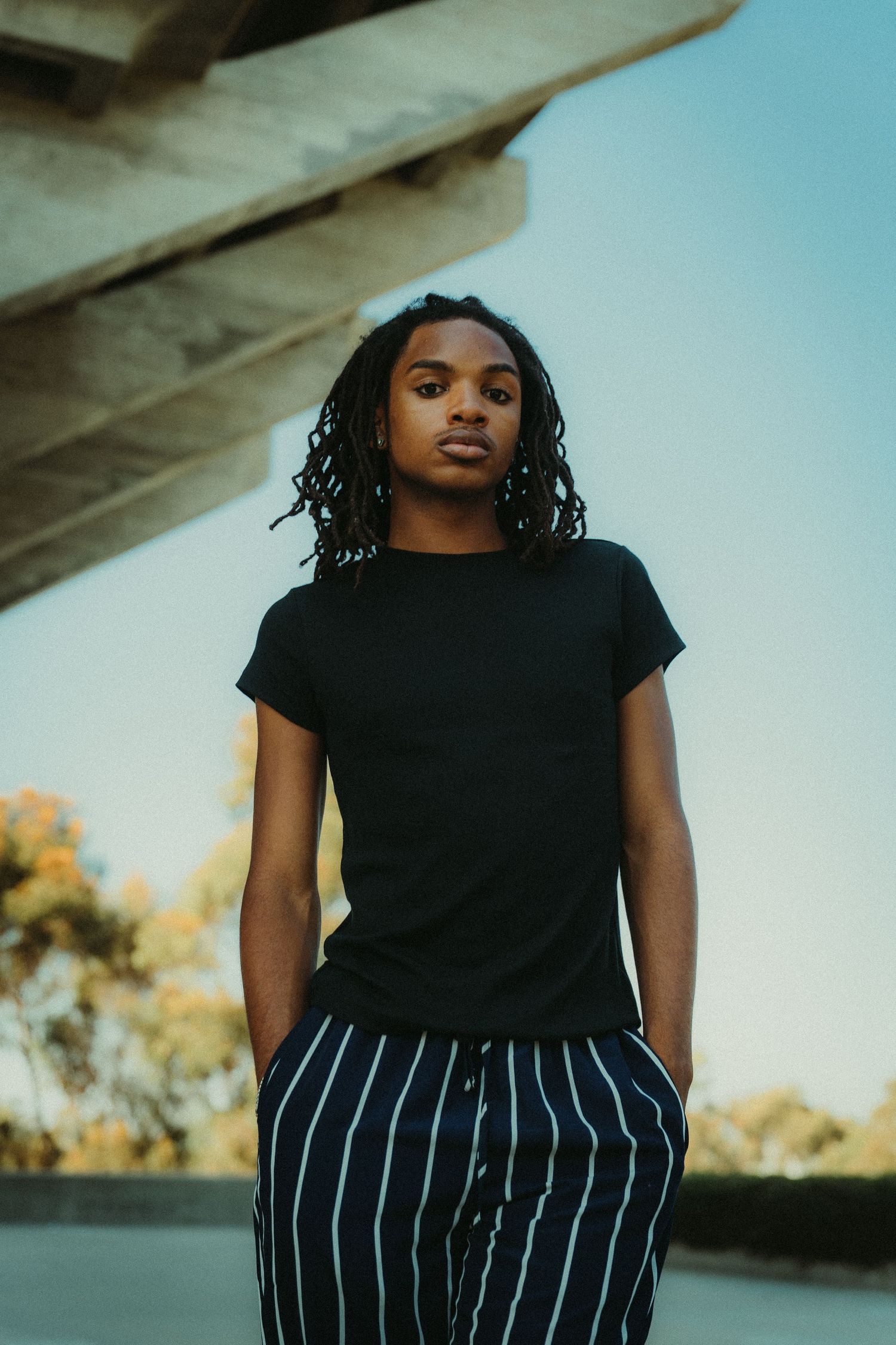 model wearing a black t-shirt and striped pants poses casually against a blurred background in warm evening light during fashion photoshoot