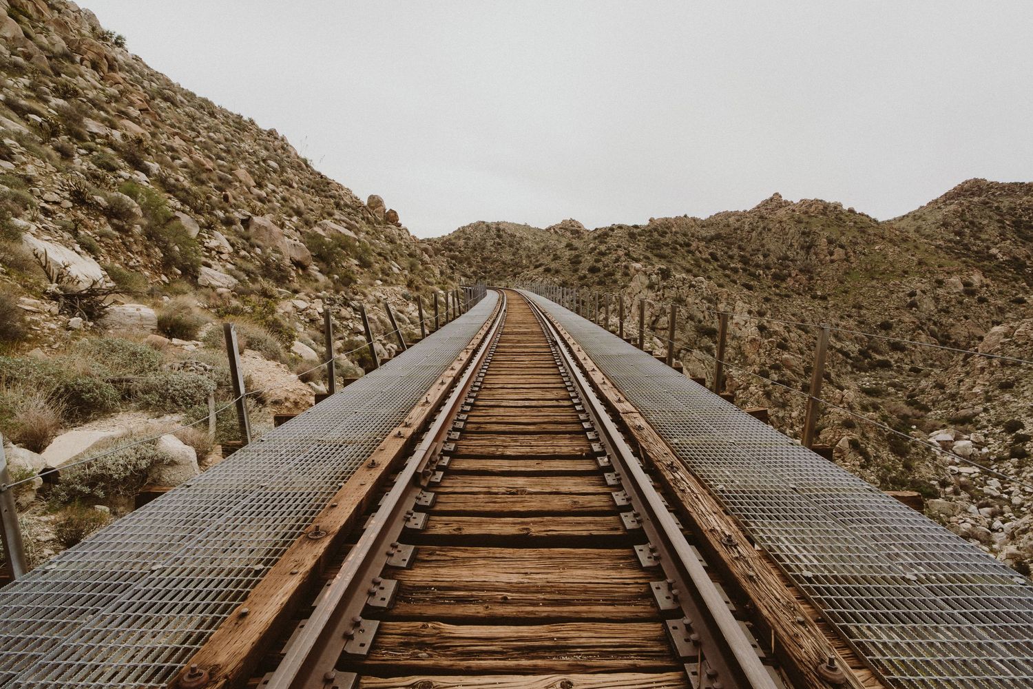Wooden railroad tracks curve through a desert mountain landscape with rugged terrain and cacti in the background.