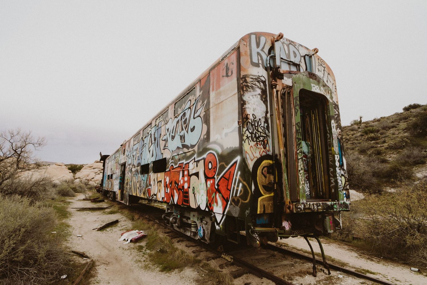 A series of abandoned train cars covered in colorful graffiti art sit along desert tracks at sunset.