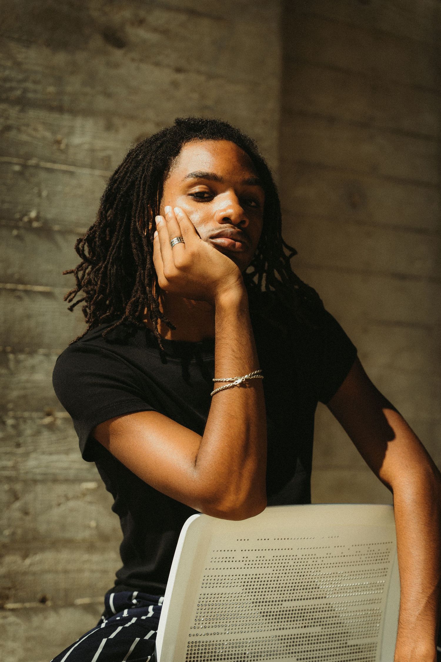 A creative portrait of someone in a black t-shirt and dreadlocks sitting pensively by a concrete wall.