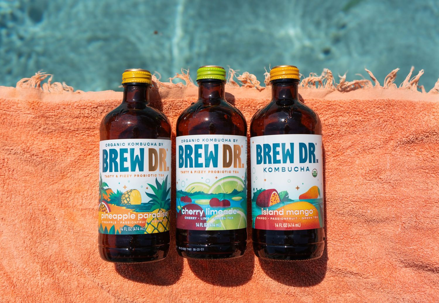 Three bottles of Brew Dr. Kombucha lined up on an orange towel by a pool.