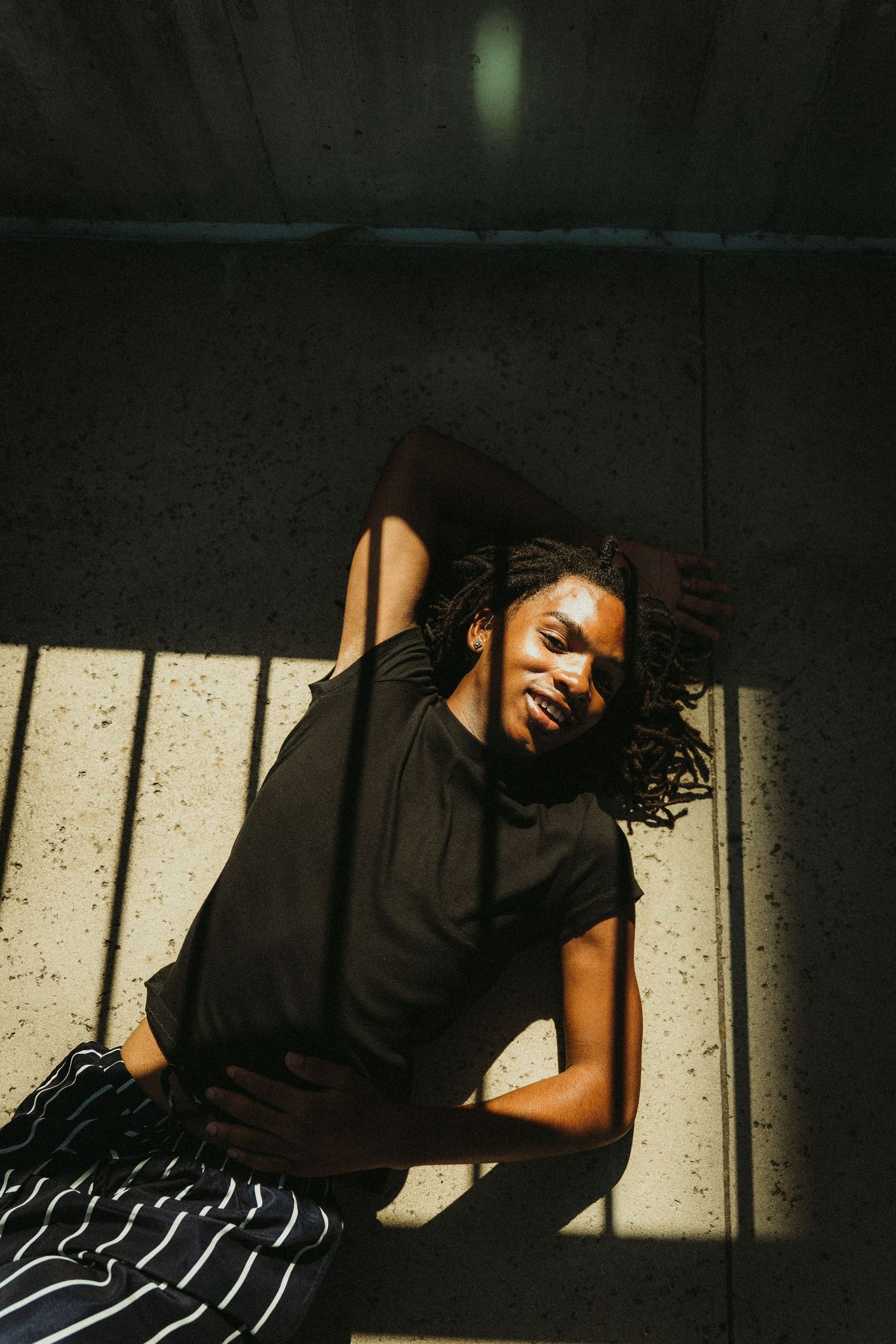 Joyful person in black top and striped pants lounging in dramatic sunlight against concrete wall during creative portrait photoshoot