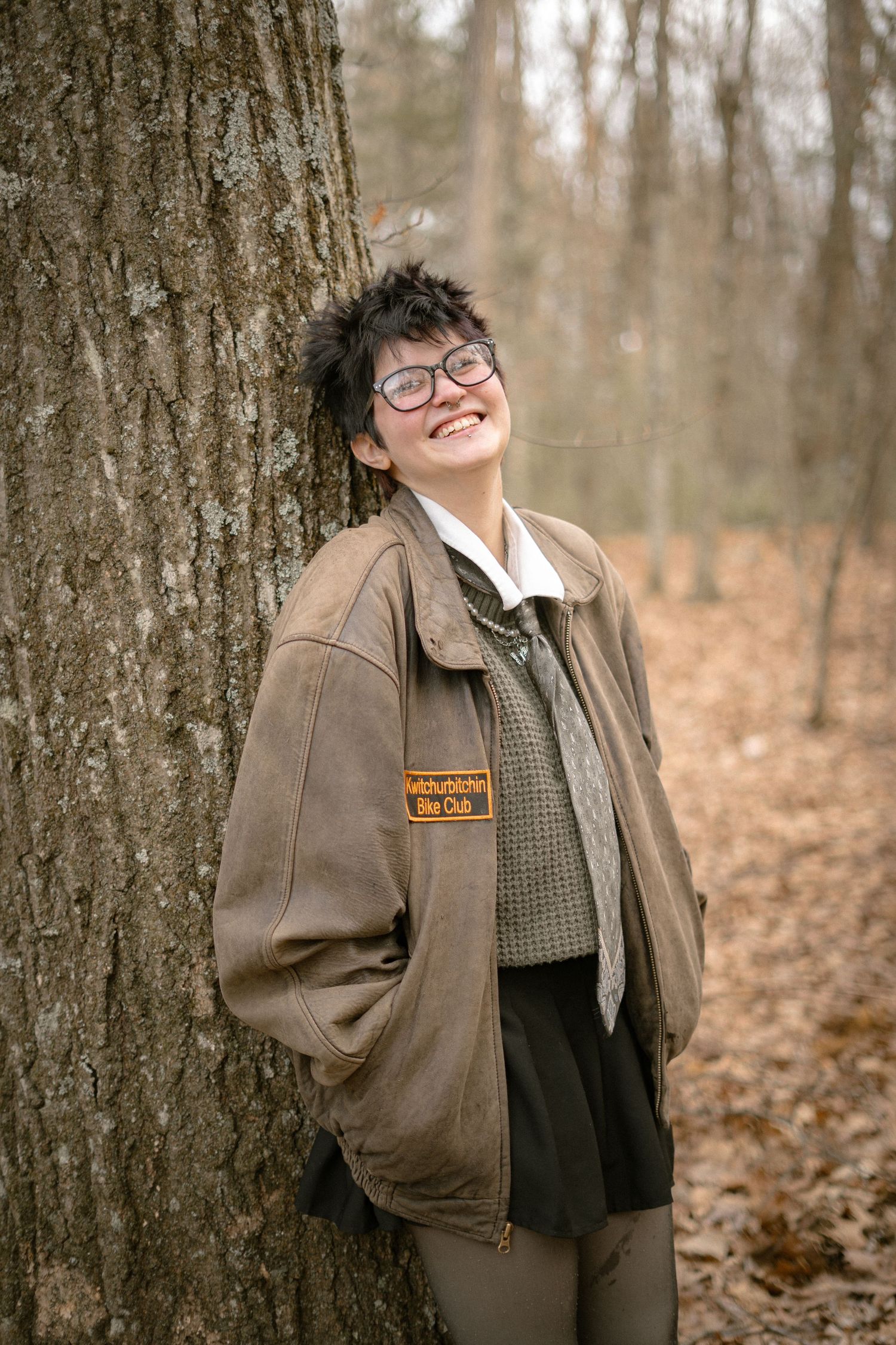 A person in a brown jacket and glasses poses casually against a tree trunk in an autumn forest setting.