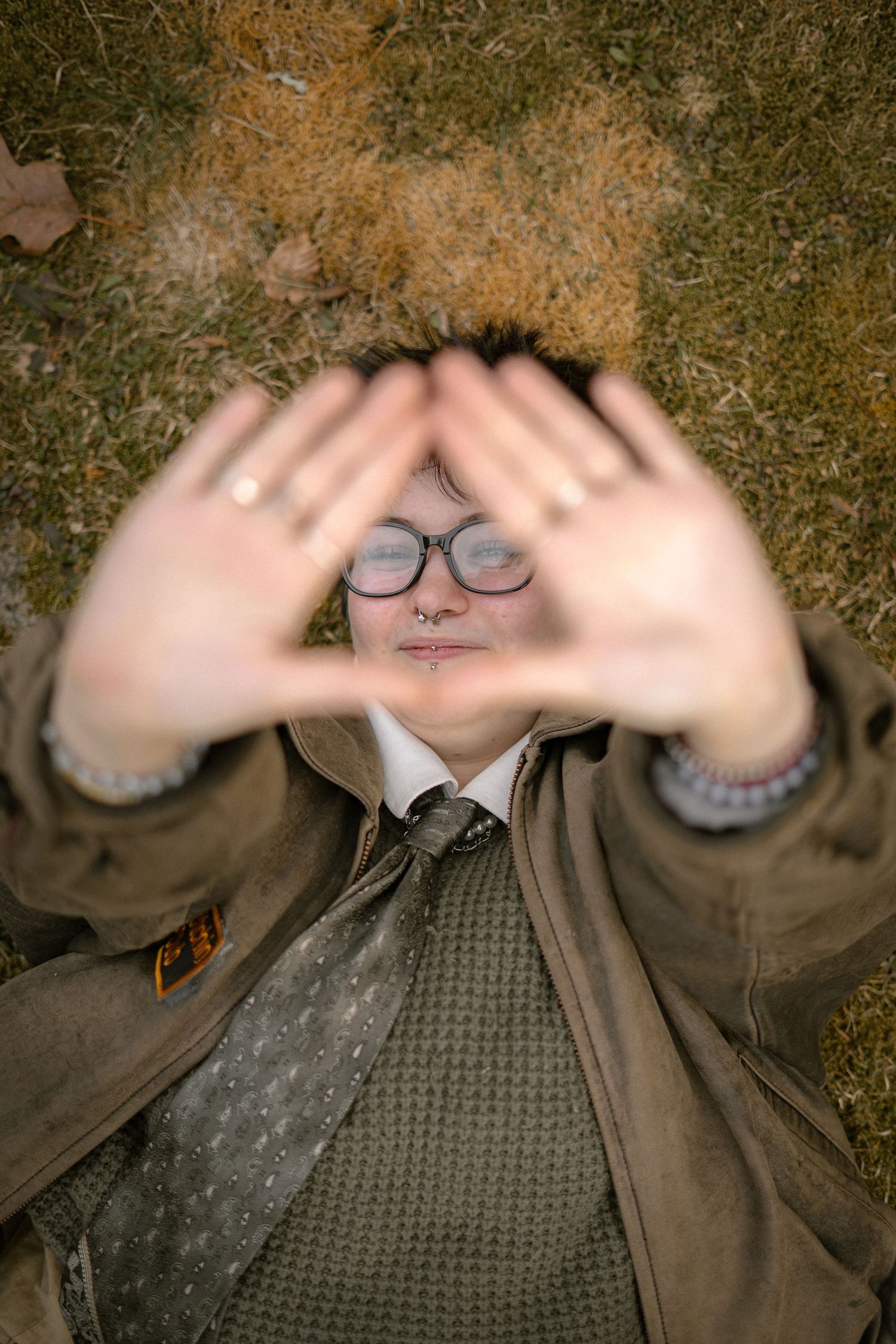 A person wearing glasses holds up their hands framing their head while lying on grass.