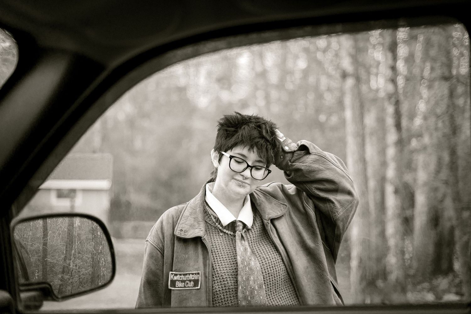 Black and white photo taken from inside a car showing person in glasses and jacket against a foggy forest backdrop.