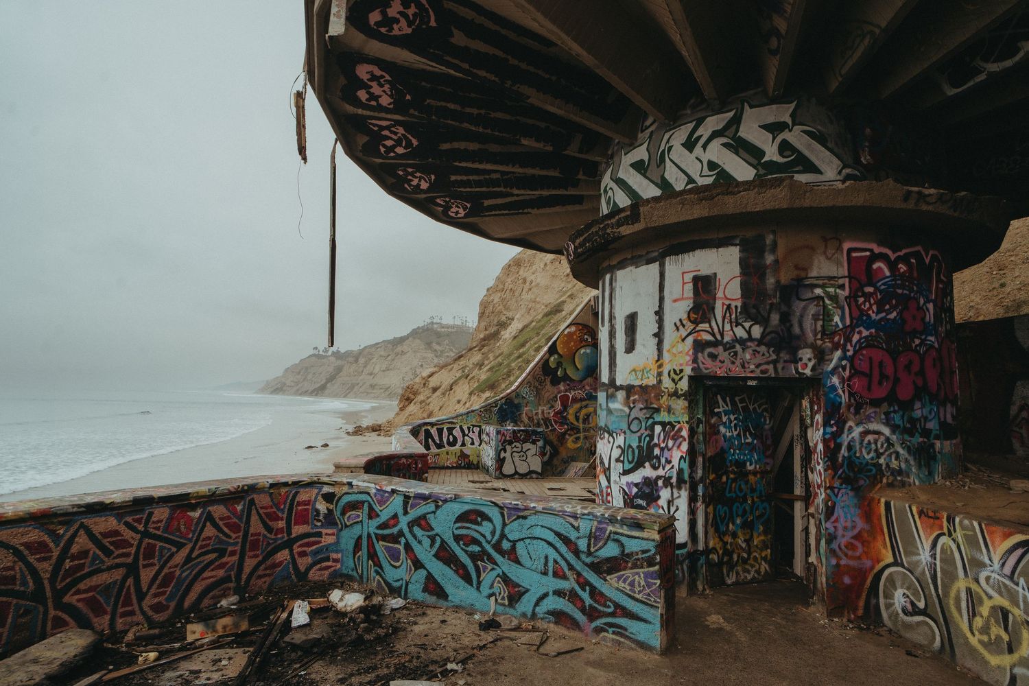 Graffiti art covers concrete pillars and walls under a bridge or overpass structure in an urban setting.