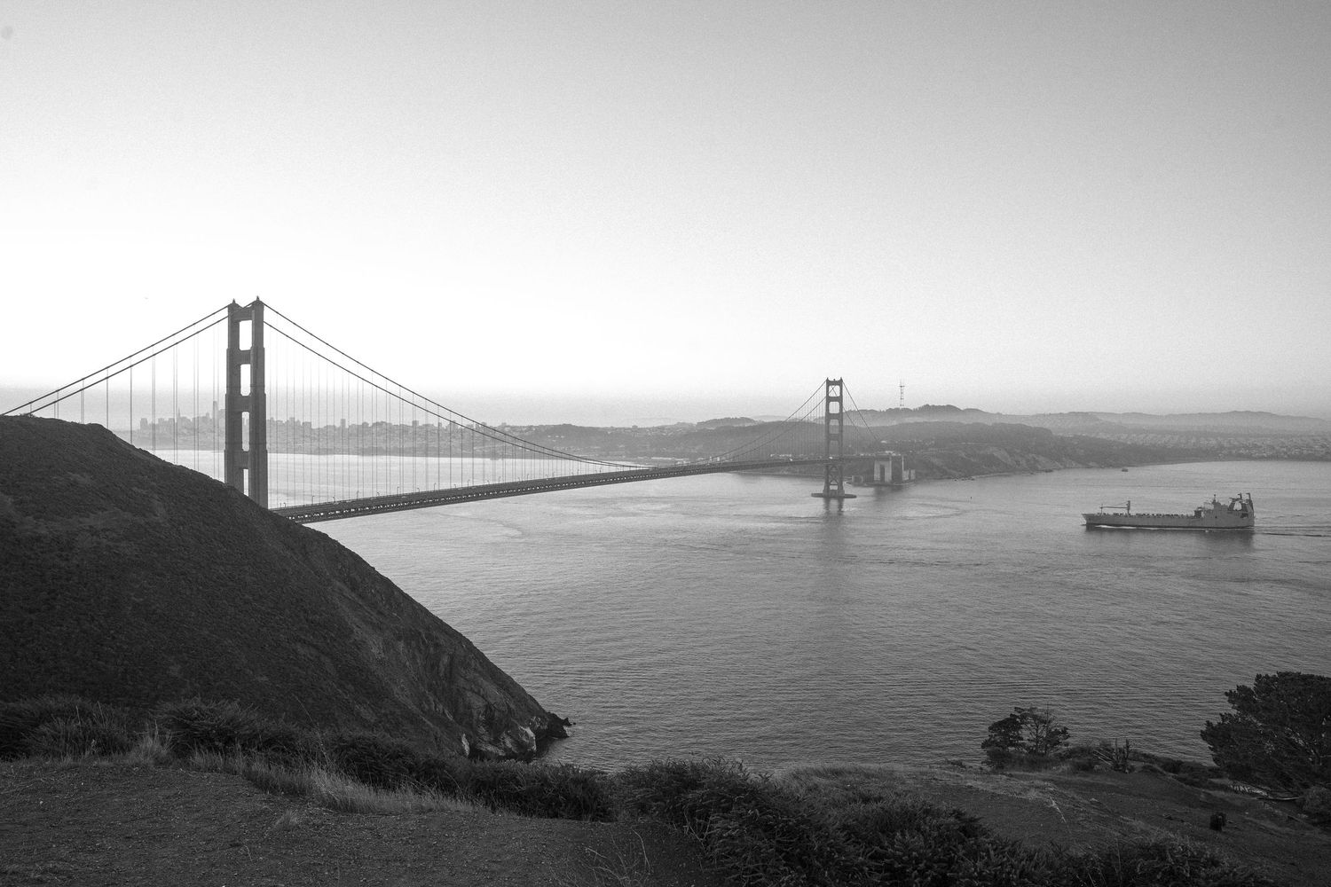 Black and white view of the Golden Gate Bridge spanning San Francisco Bay with a ship passing underneath in misty conditions.
