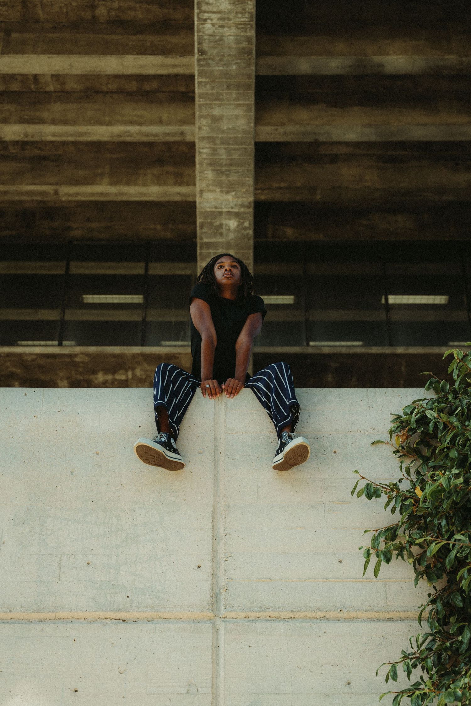 model in black shirt sits on a white ledge against a wooden wall background with greenery nearby during lifestyle photoshoot