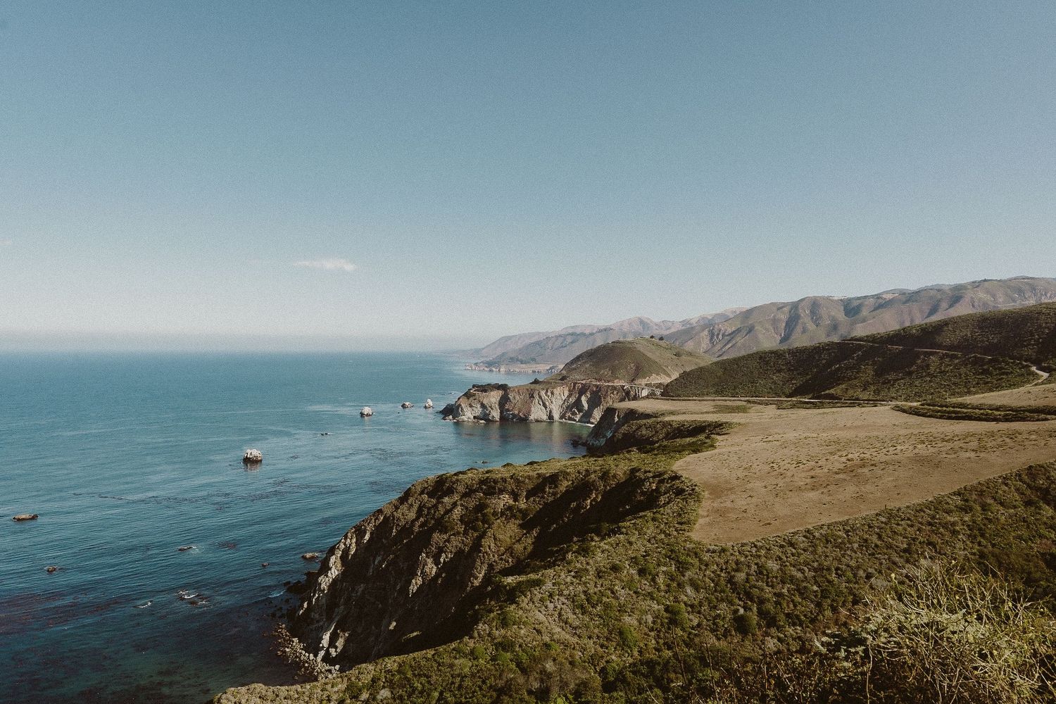 Scenic aerial views of rugged coastal cliffs with turquoise ocean waters and winding paths along the Pacific Coast Highway.
