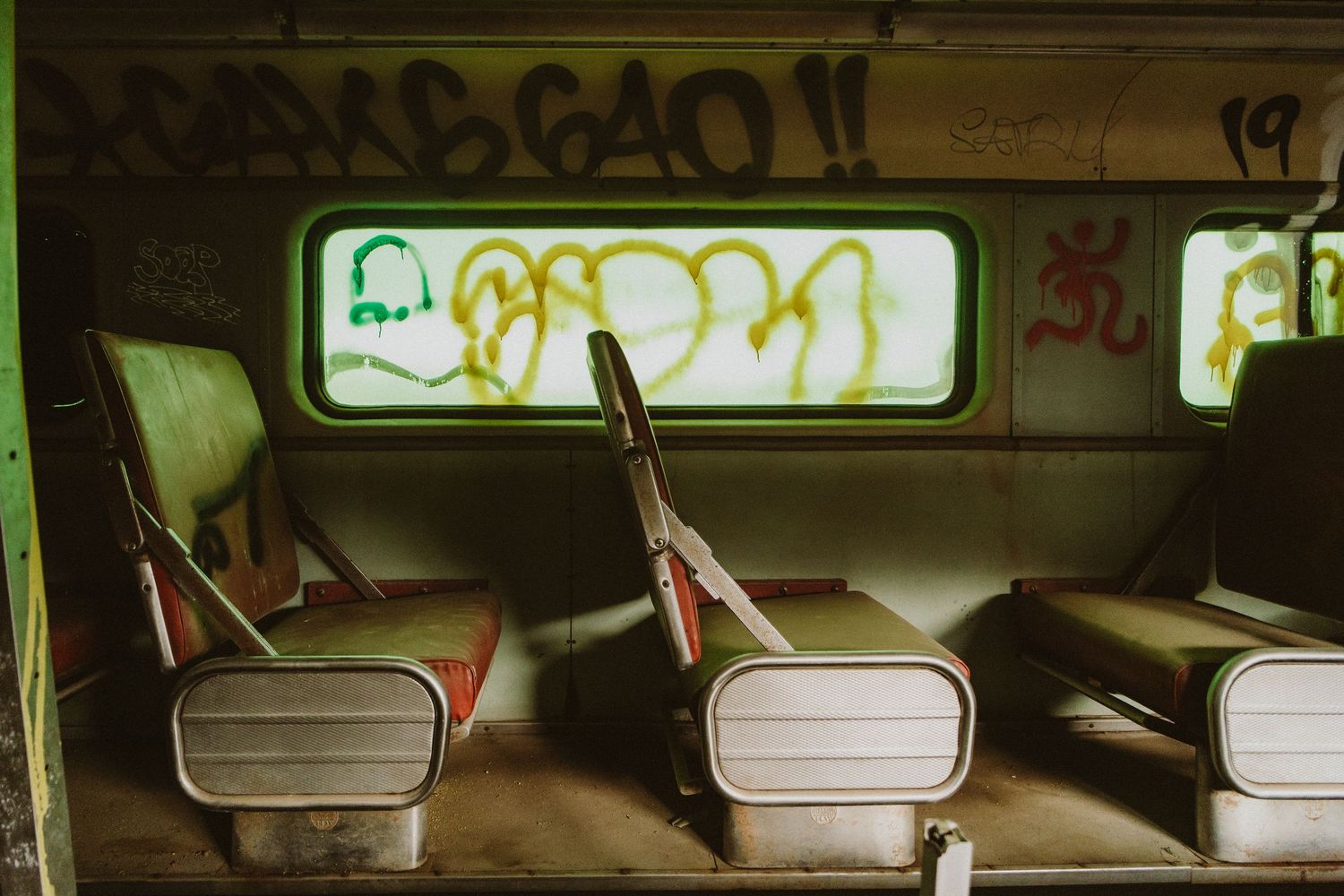 Vintage subway car interior showing worn leather seats and graffiti-covered windows with green lighting.