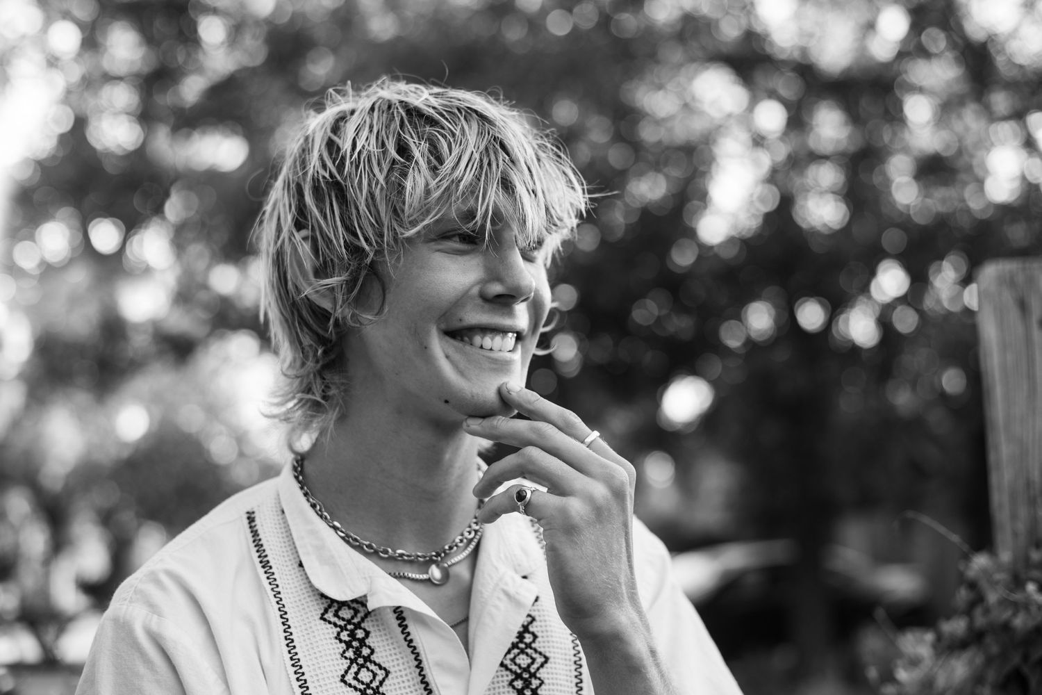 Black and white portrait of a brand photographer with blonde hair smiling while touching their chin in outdoor setting.