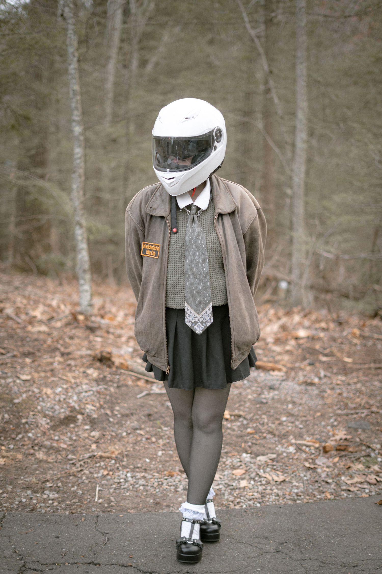 Figure in vintage-style outfit with leather jacket, gray skirt and white motorcycle helmet poses in wooded area.