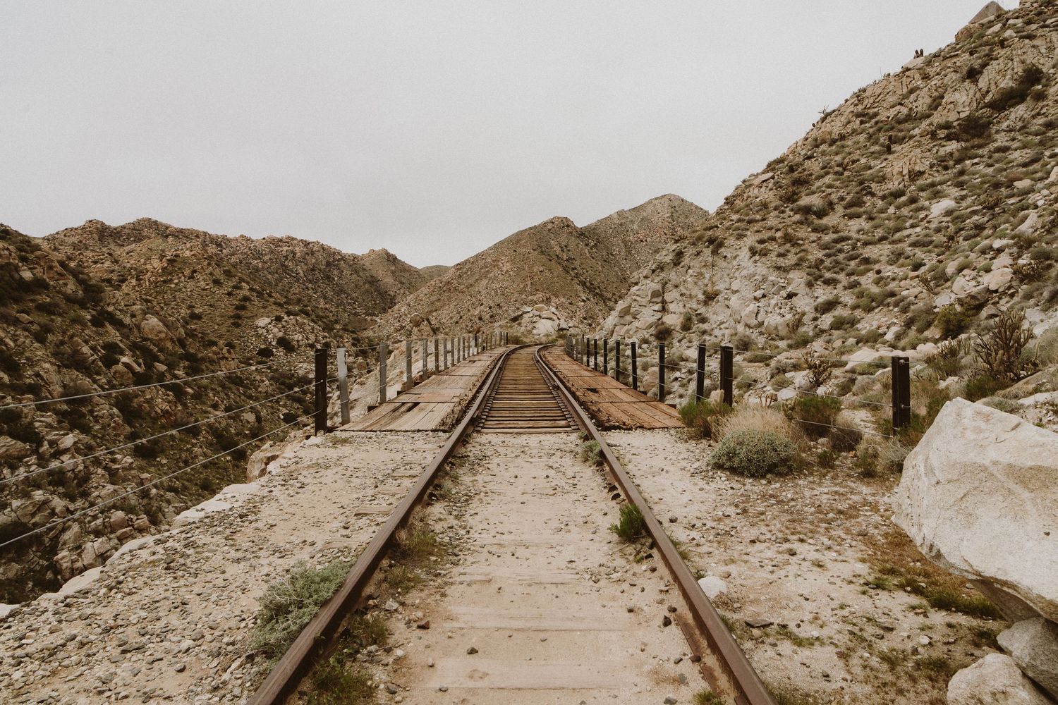 Vintage sepia-toned railroad tracks leading through rugged desert mountains with wooden posts along both sides.
