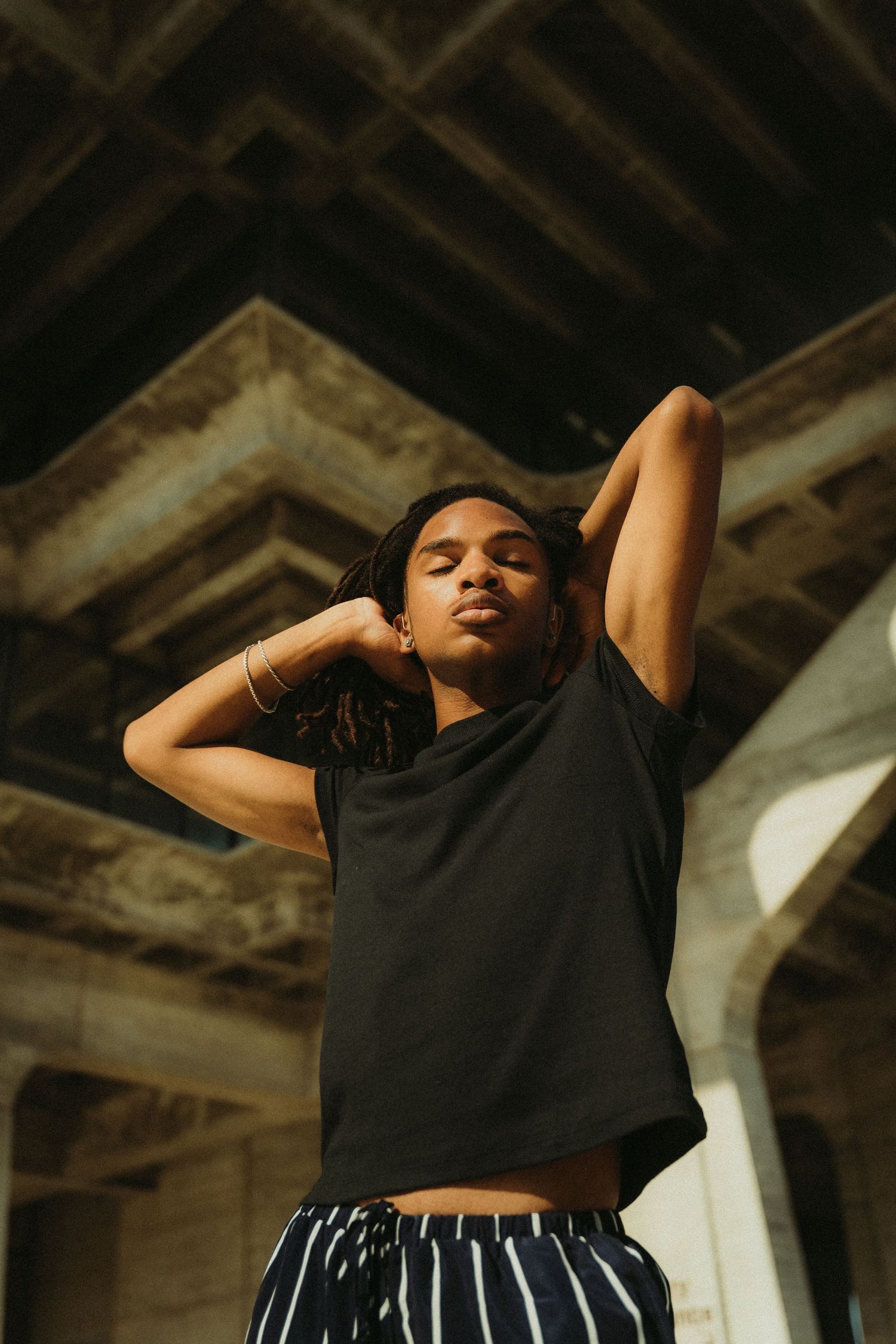 A model in a black t-shirt and striped pants stretches with hands behind their head under an architectural structure during a portrait shoot