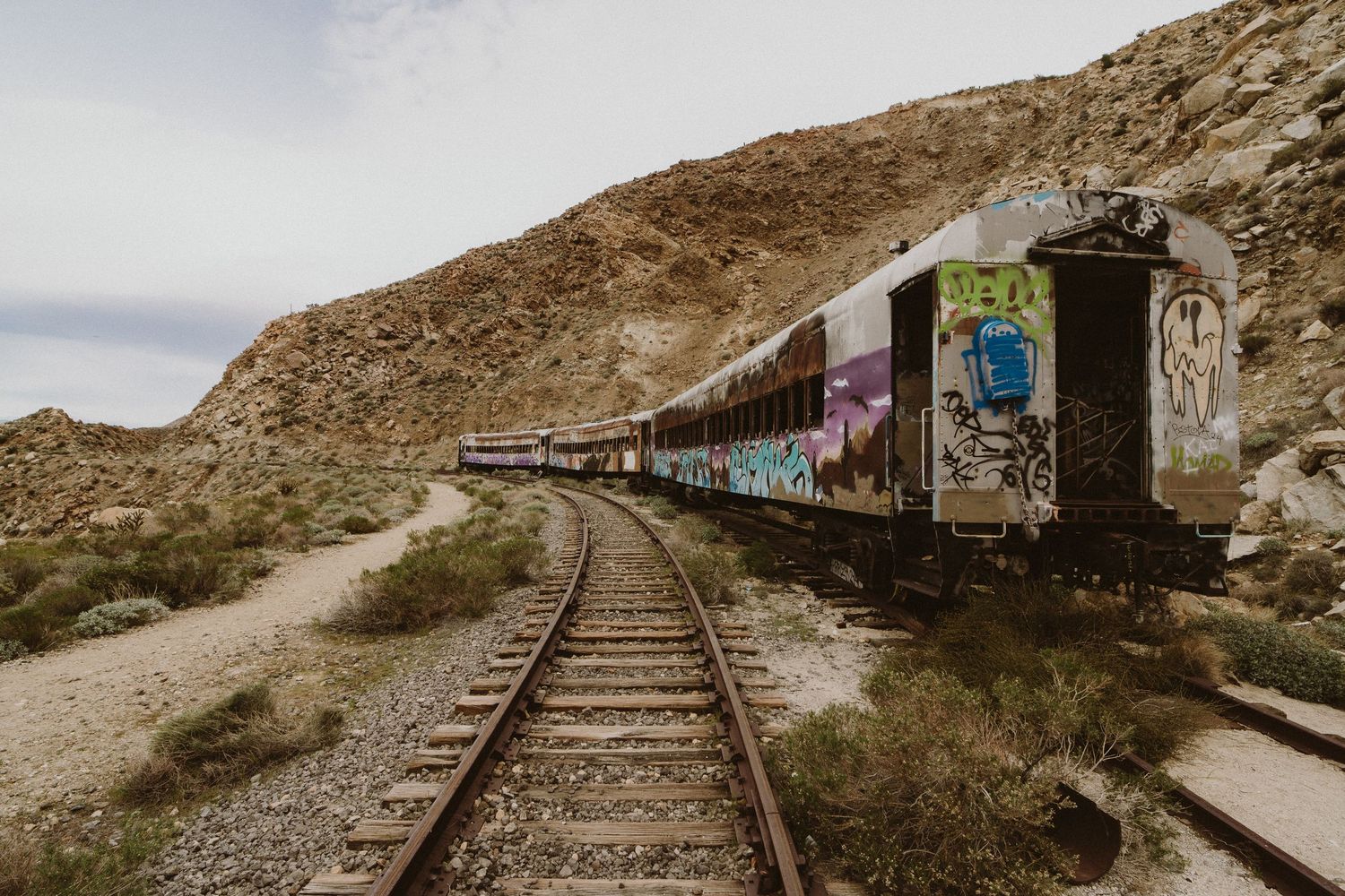 Abandoned graffiti-covered train car sits on curved railroad tracks through desert landscape with rocky hills in background.