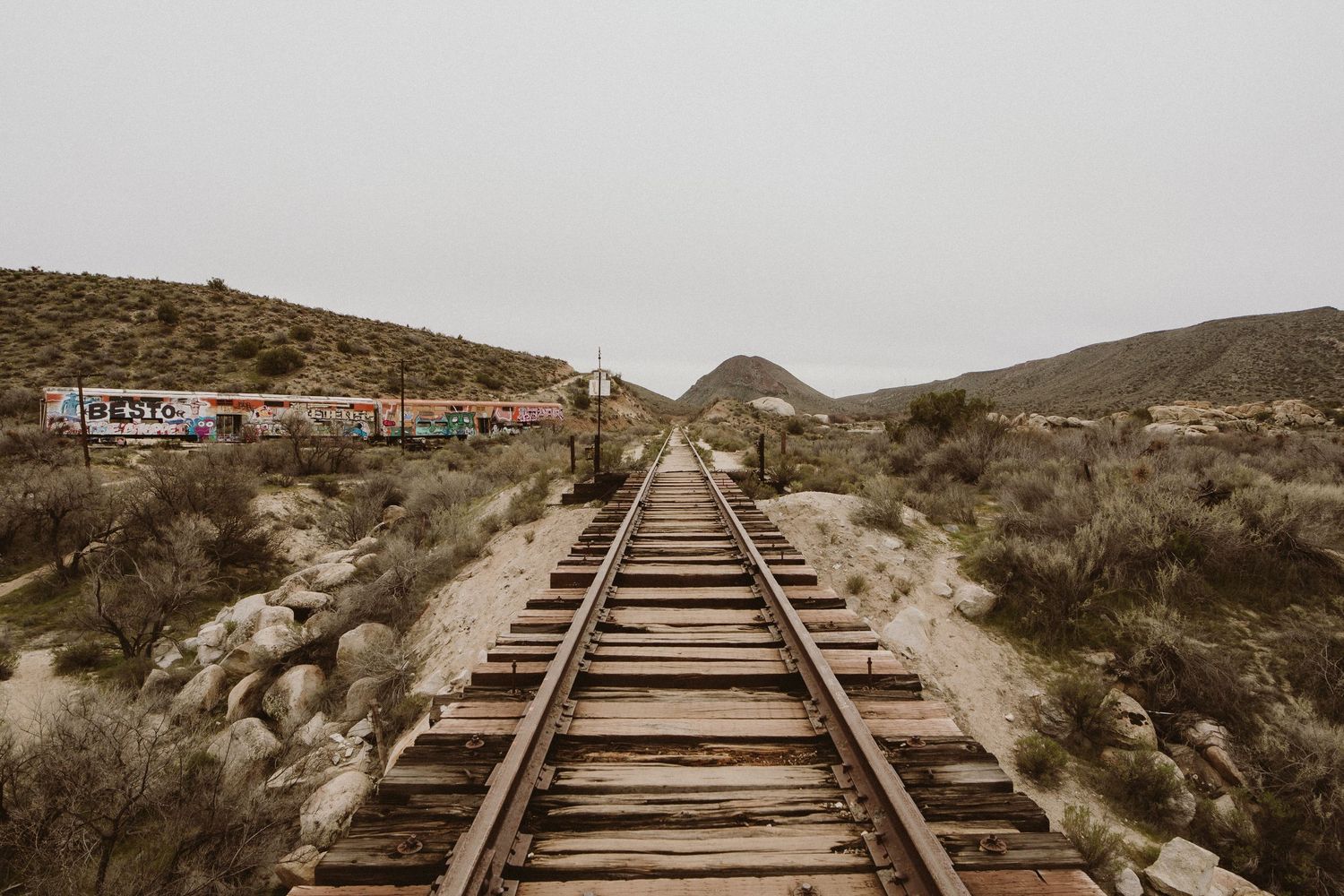Old wooden railroad tracks leading through a desert landscape with hills and scrub brush in the background.