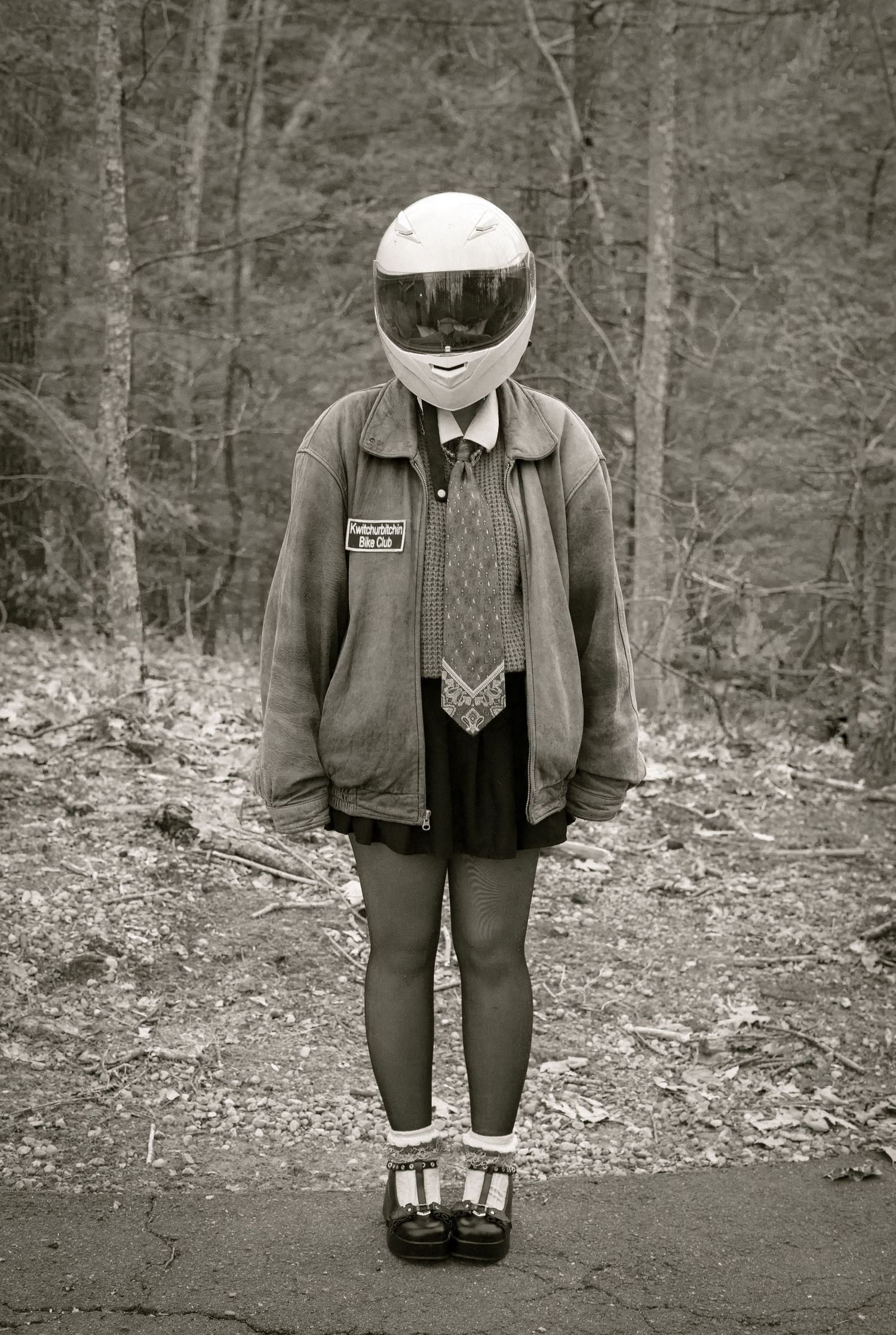 A figure wearing a motorcycle helmet, jacket and dress stands in a wooded area in black and white photography.