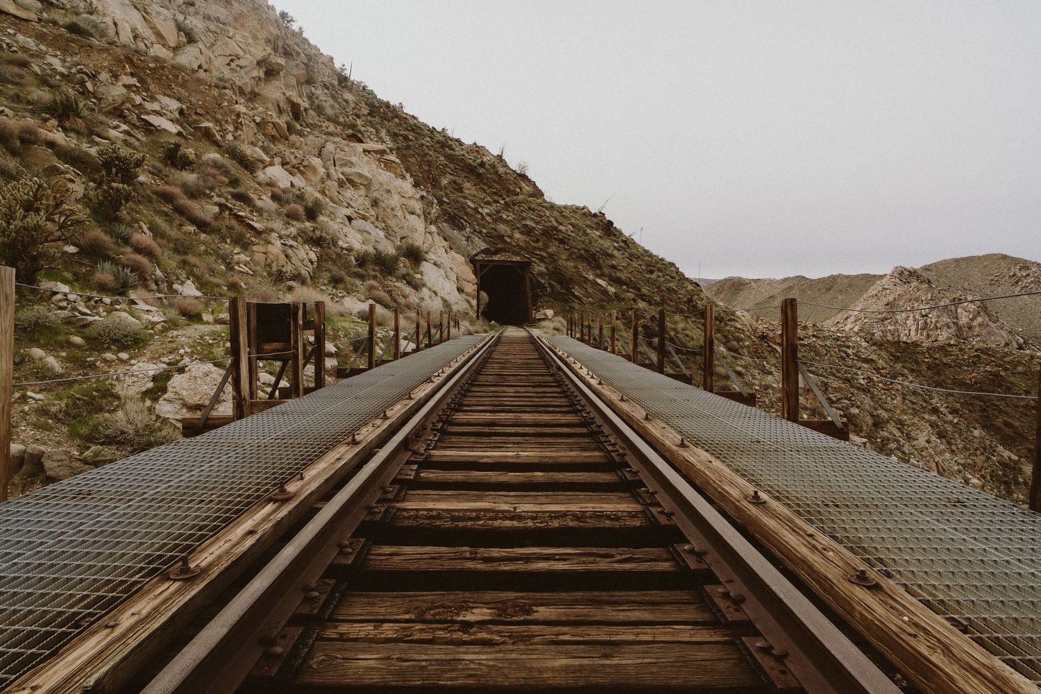 Historic wooden railroad trestle bridge spans across rugged mountain terrain in a desert landscape.