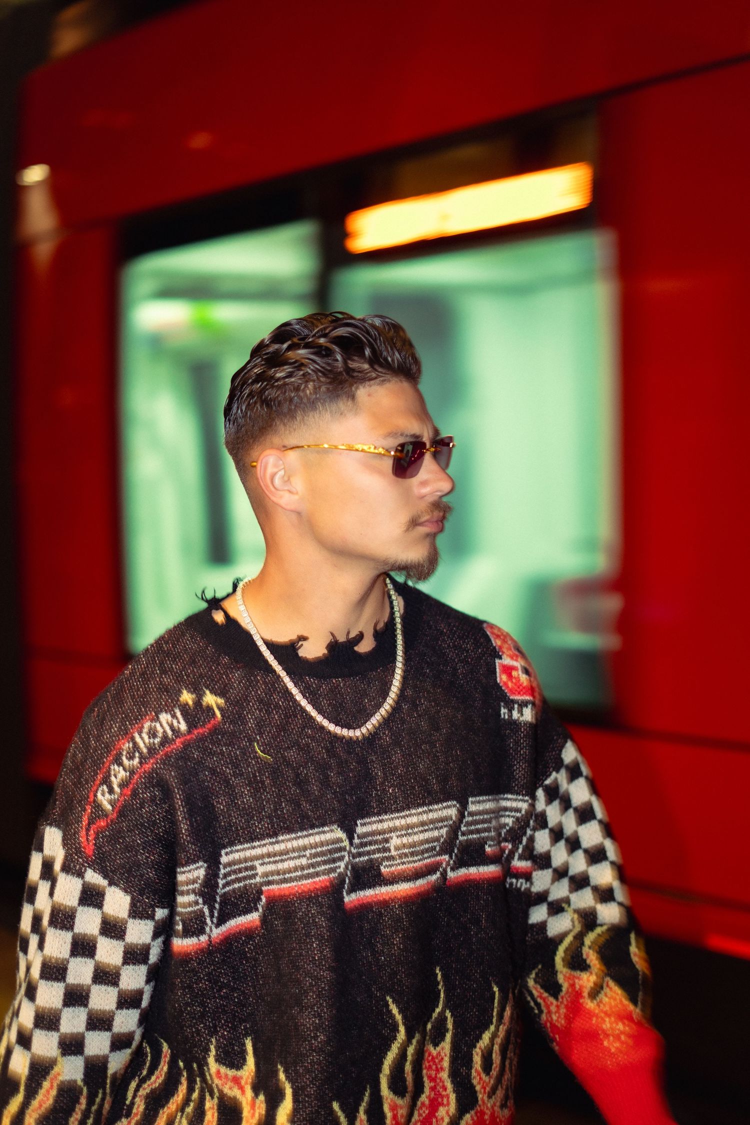 Street style portrait featuring flame pattern sweater and sunglasses against red transit background.