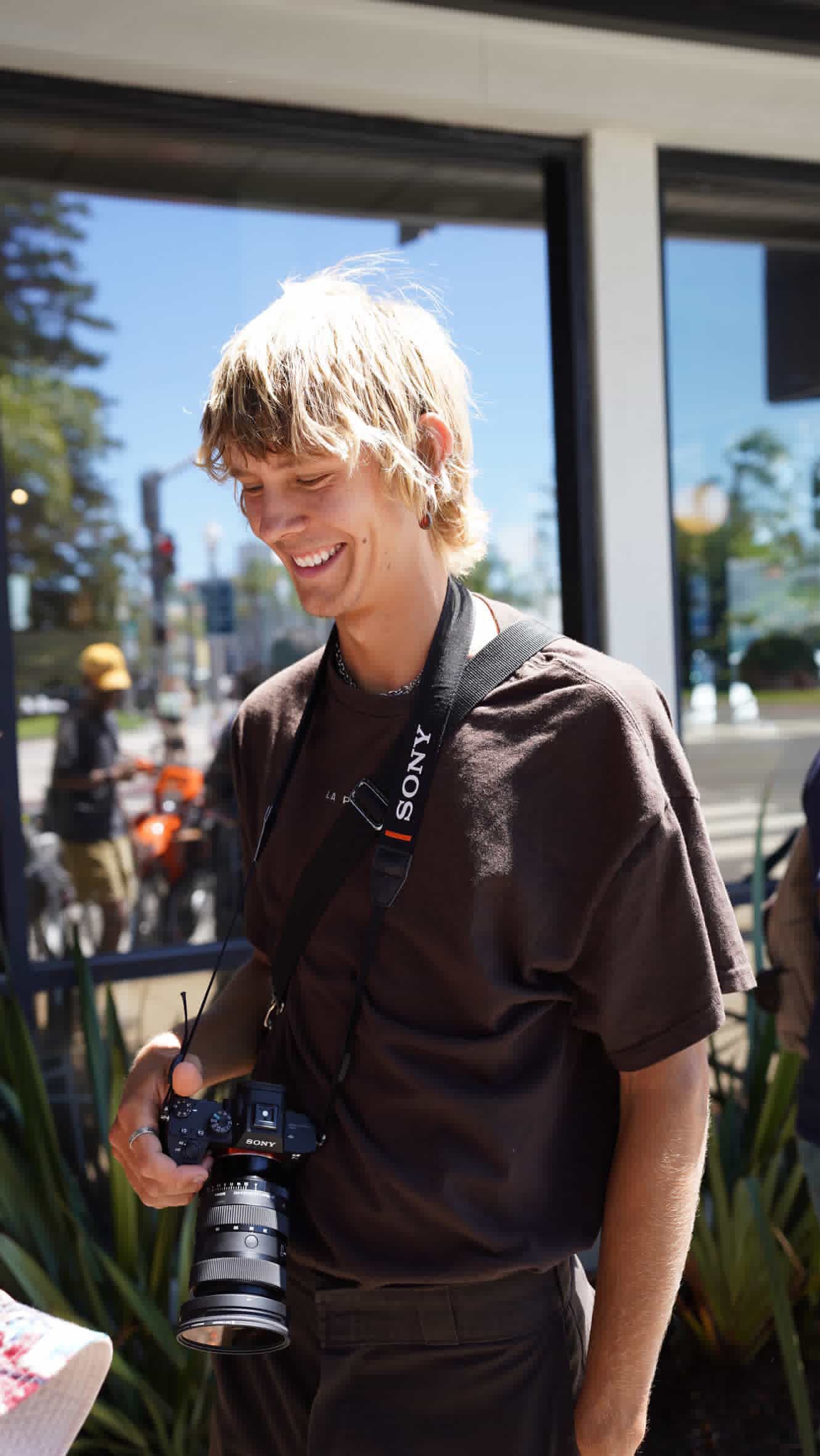 A person in a black shirt smiles and holds a camera while standing outdoors on a sunny day.