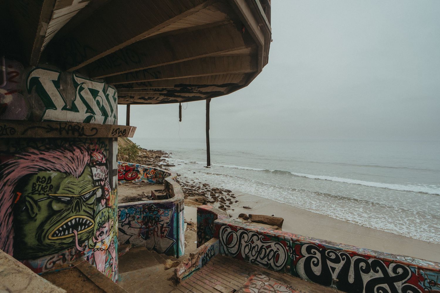 Graffiti-covered concrete walls line a coastal pathway with crashing ocean waves visible in the distance.
