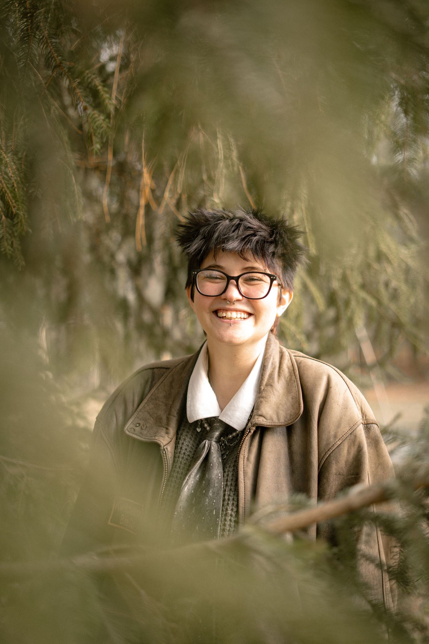 Portrait through foliage showing someone wearing glasses and brown jacket while smiling warmly outdoors.