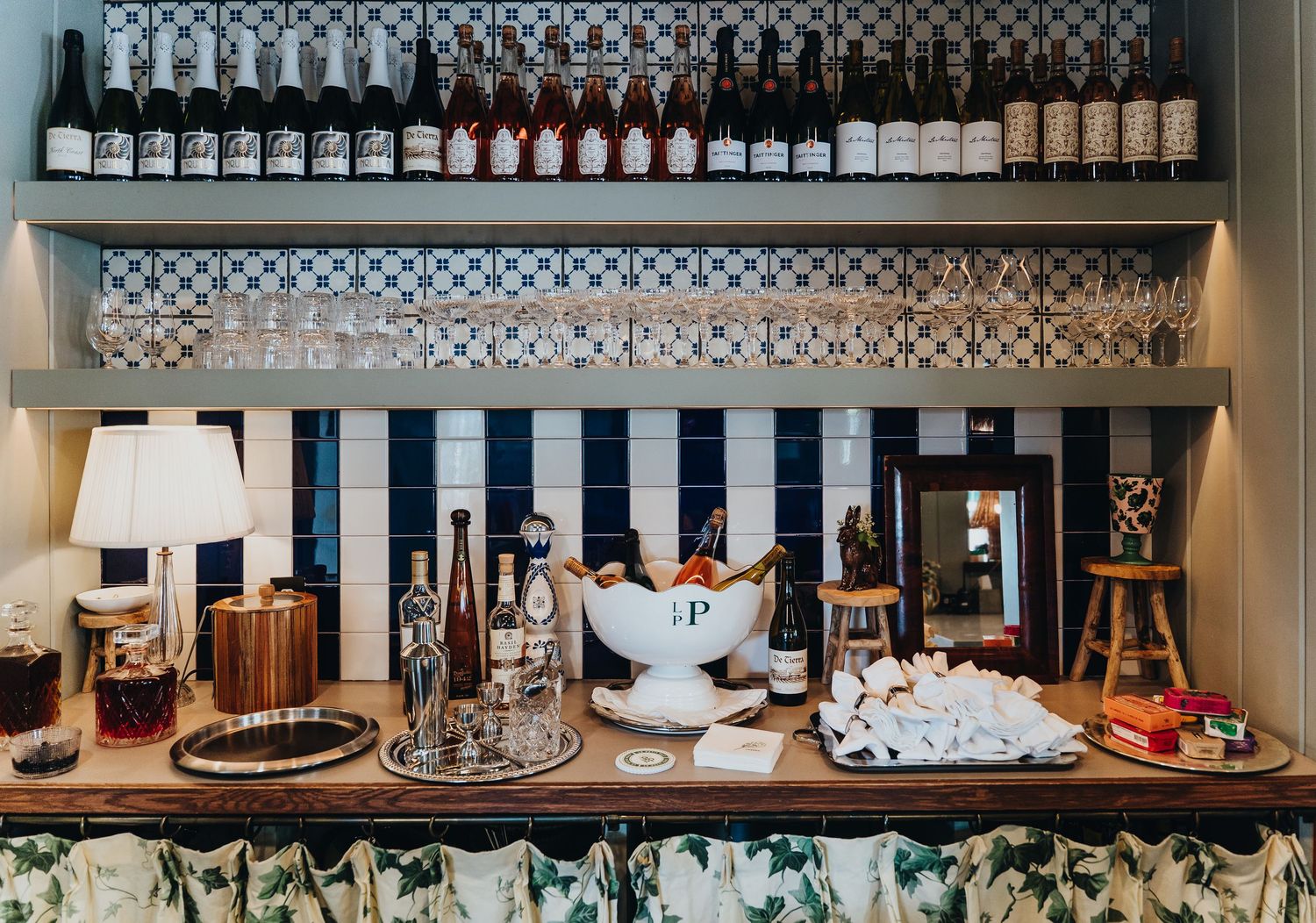 Long shelf display of neatly arranged liquor bottles and glassware in a bar or restaurant setting.