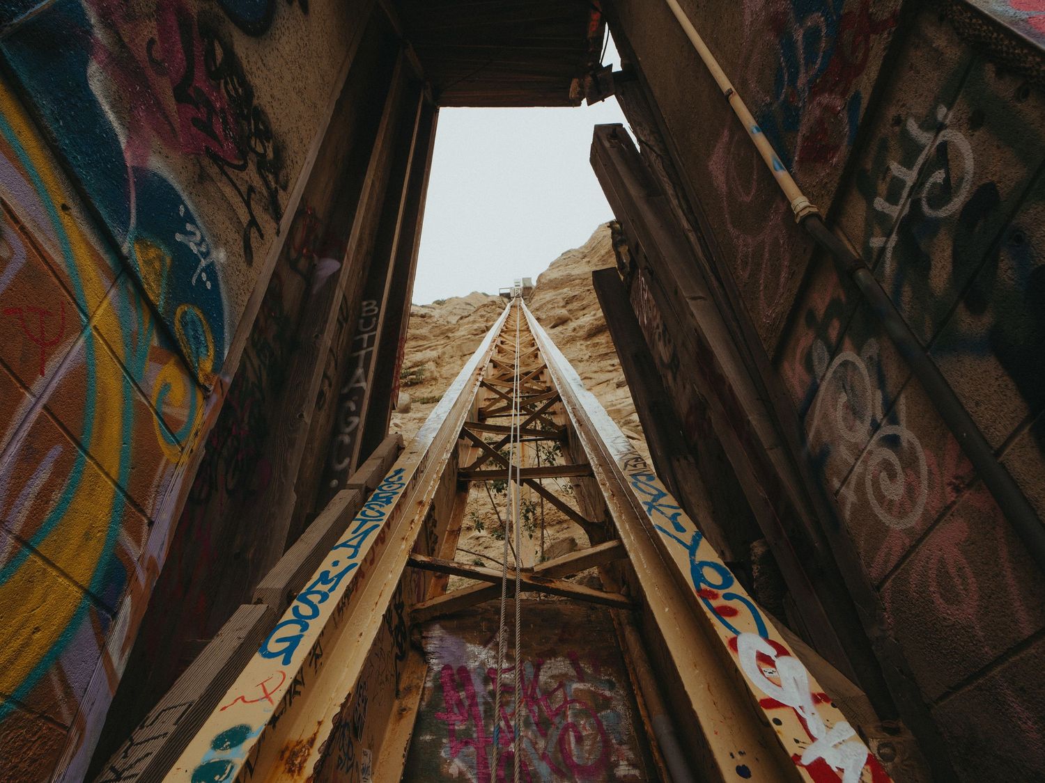 Looking up through a triangular wooden structure covered in colorful graffiti art toward a bright sky.