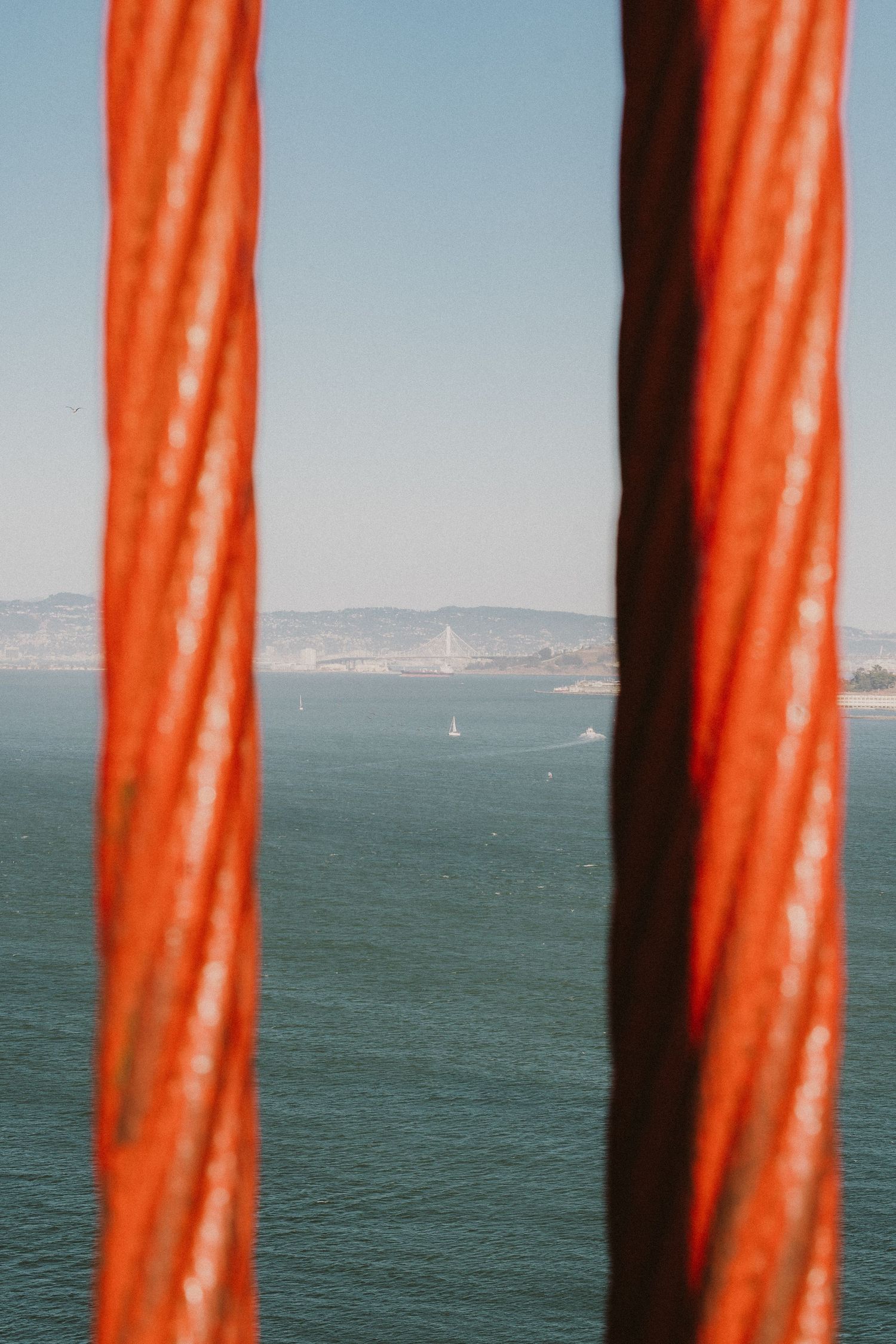 View of San Francisco Bay and distant sailboats through the iconic orange cables of the Golden Gate Bridge.