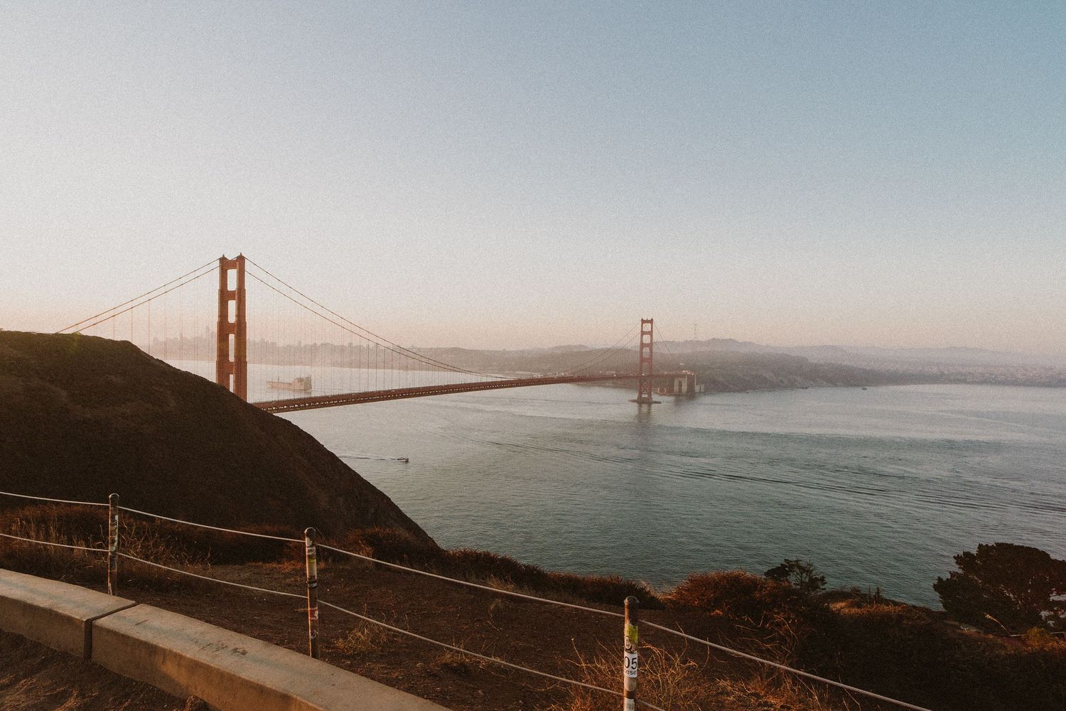 A panoramic view of the Golden Gate Bridge stretching across San Francisco Bay during sunset with coastal hills visible.