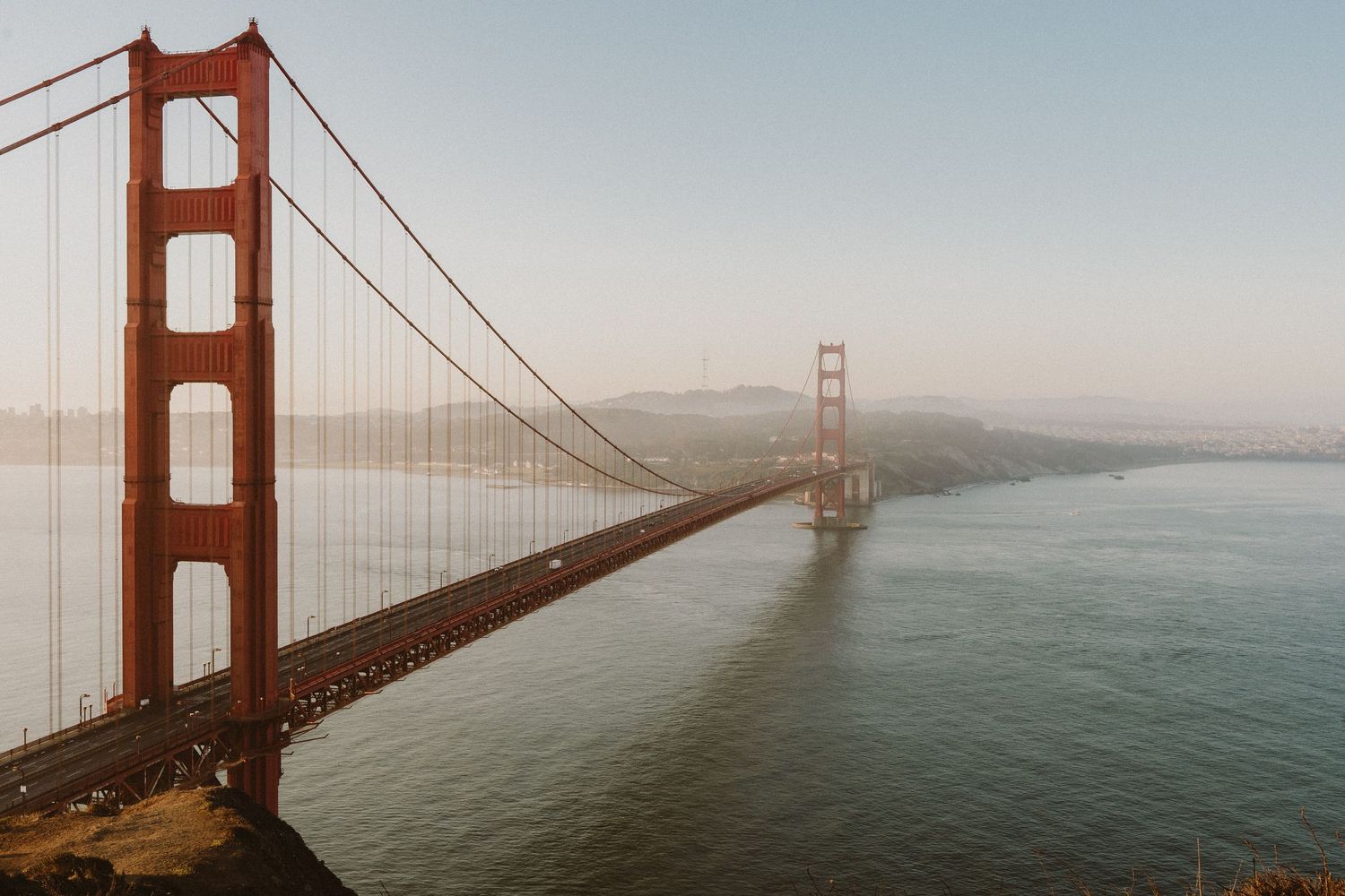 Iconic Golden Gate Bridge spans over San Francisco Bay during a misty sunset with water and mountains in background.