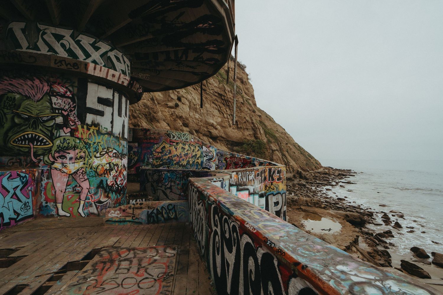 Coastal cliffs covered in graffiti art along a rocky beach with waves crashing against the shore during overcast weather.