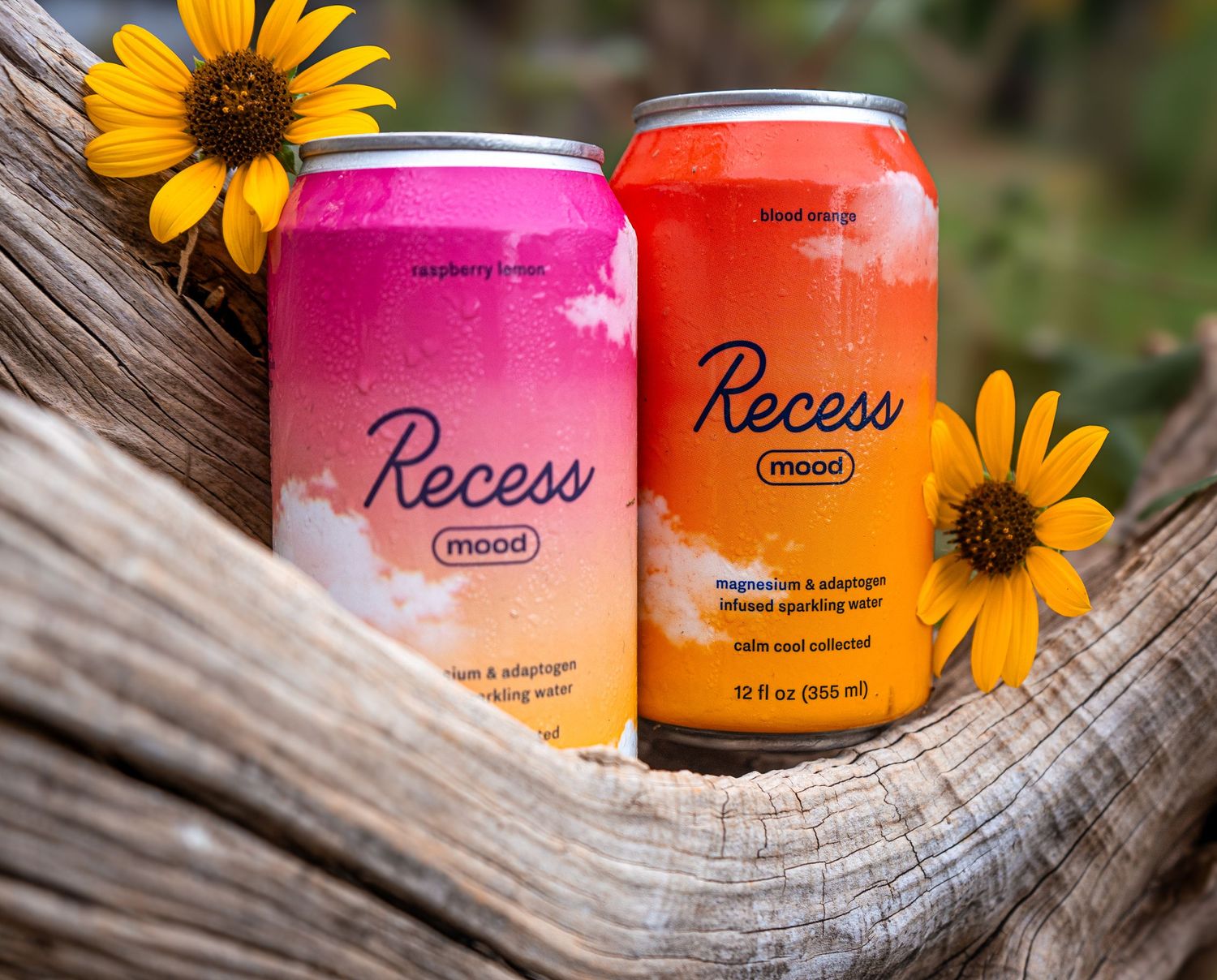 Pink and orange Recess drink cans styled with bright yellow sunflowers on wooden surface.