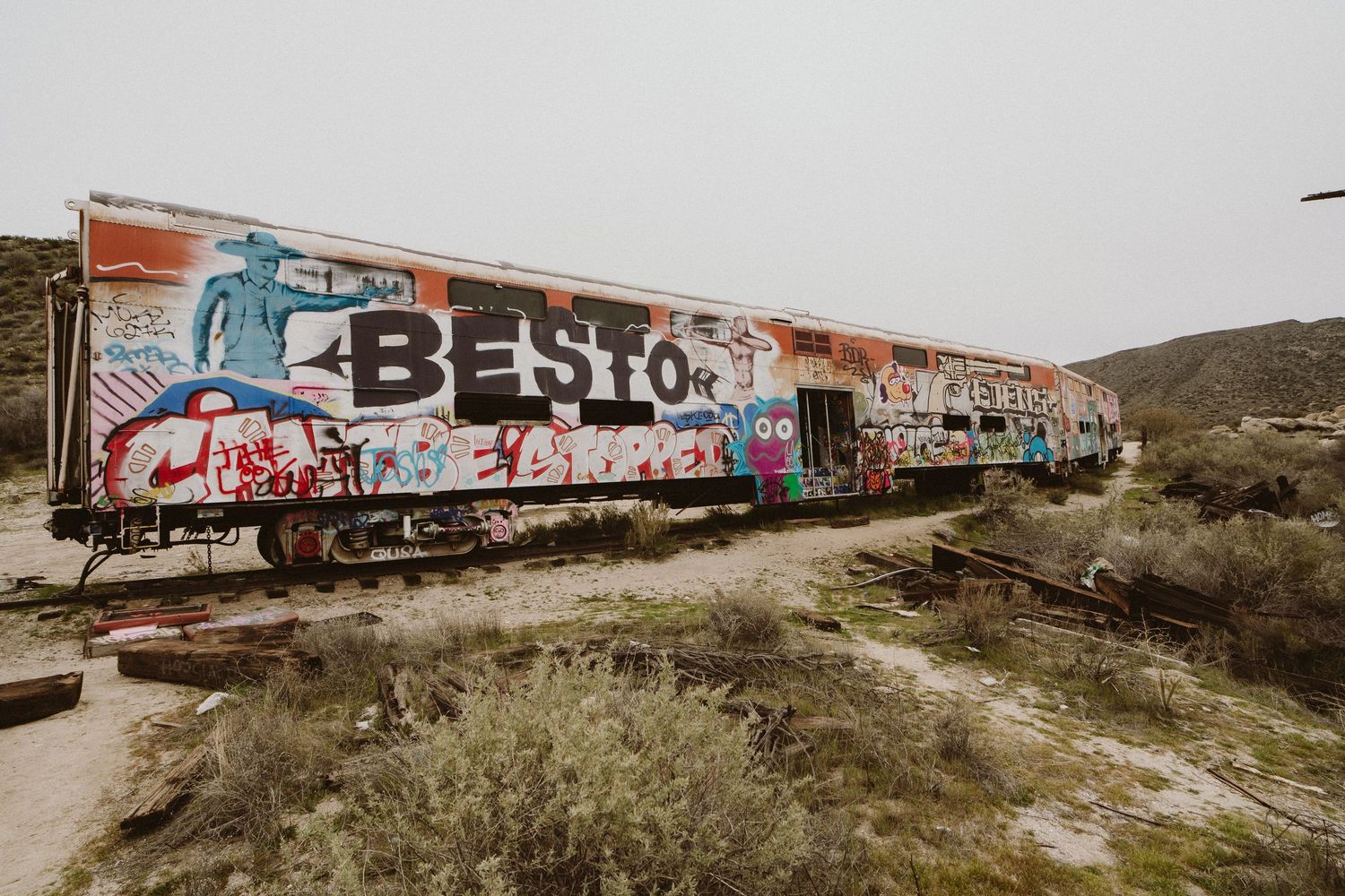 Abandoned train cars covered in colorful graffiti sit in a desert landscape with mountains in the background.