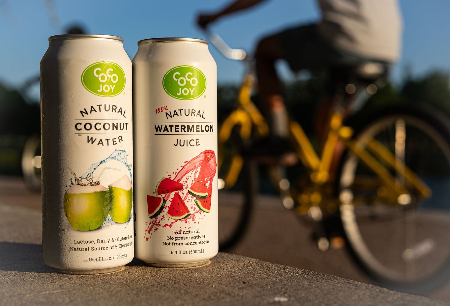 Two Coco Joy beverage cans displayed outdoors with a bicycle silhouette in the background.
