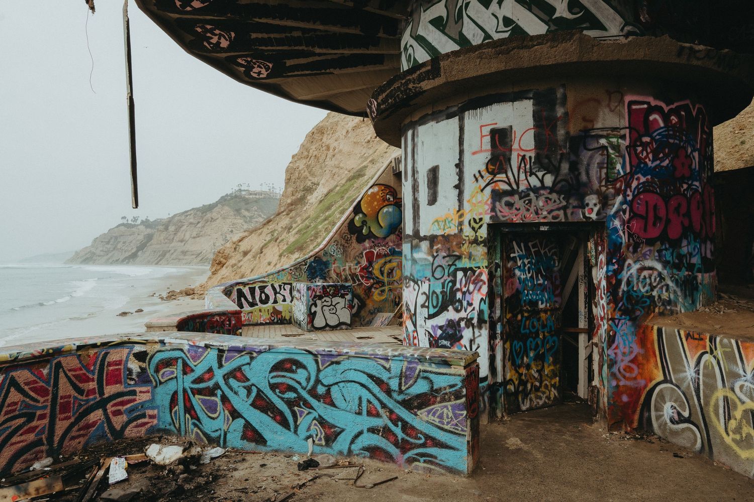 Graffiti-covered walls line a coastal tunnel pathway with ocean views and cliffs visible in the distance.