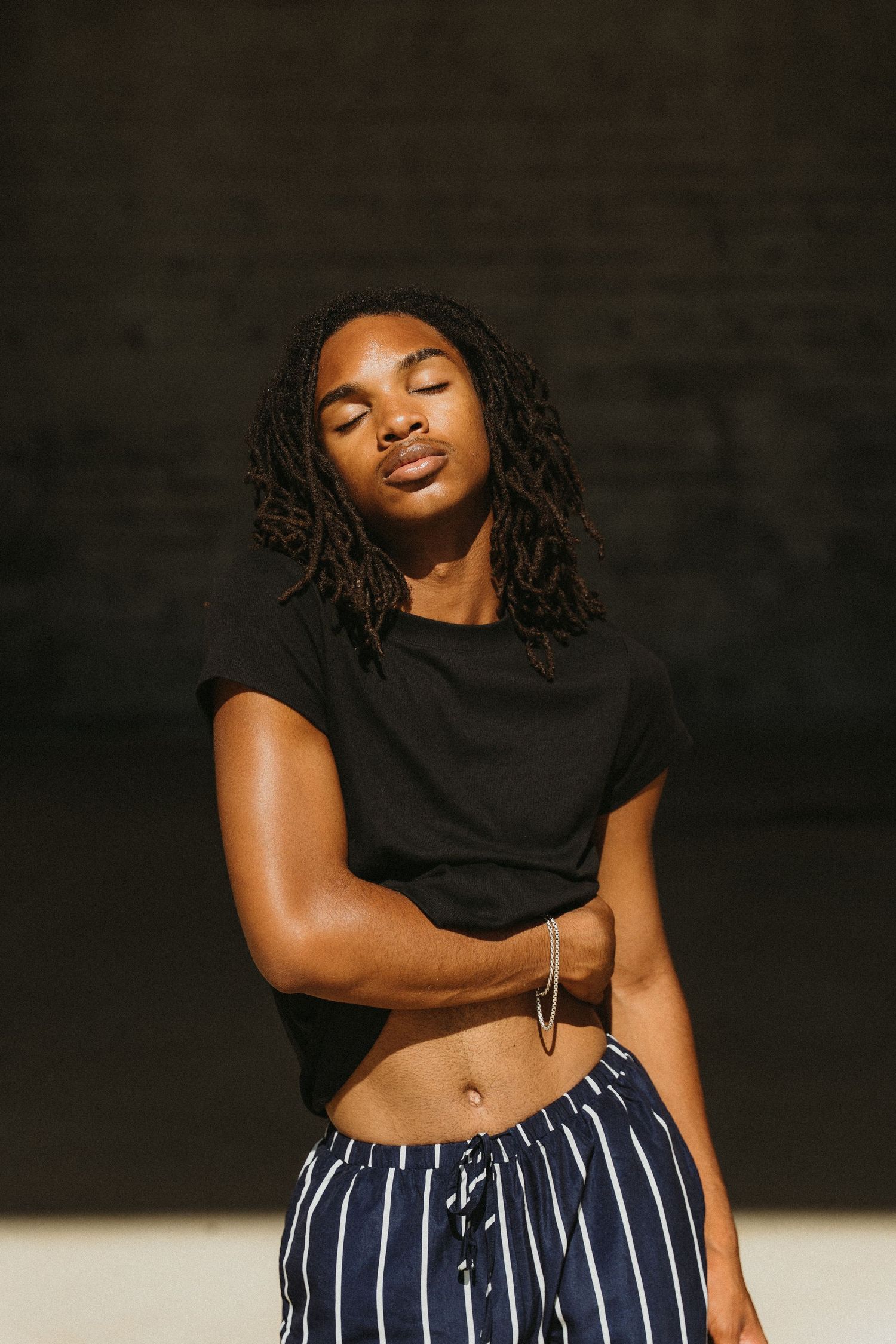 Person with dreadlocks wearing a black crop top and striped pants poses against dark background in warm lighting for brand photoshoot