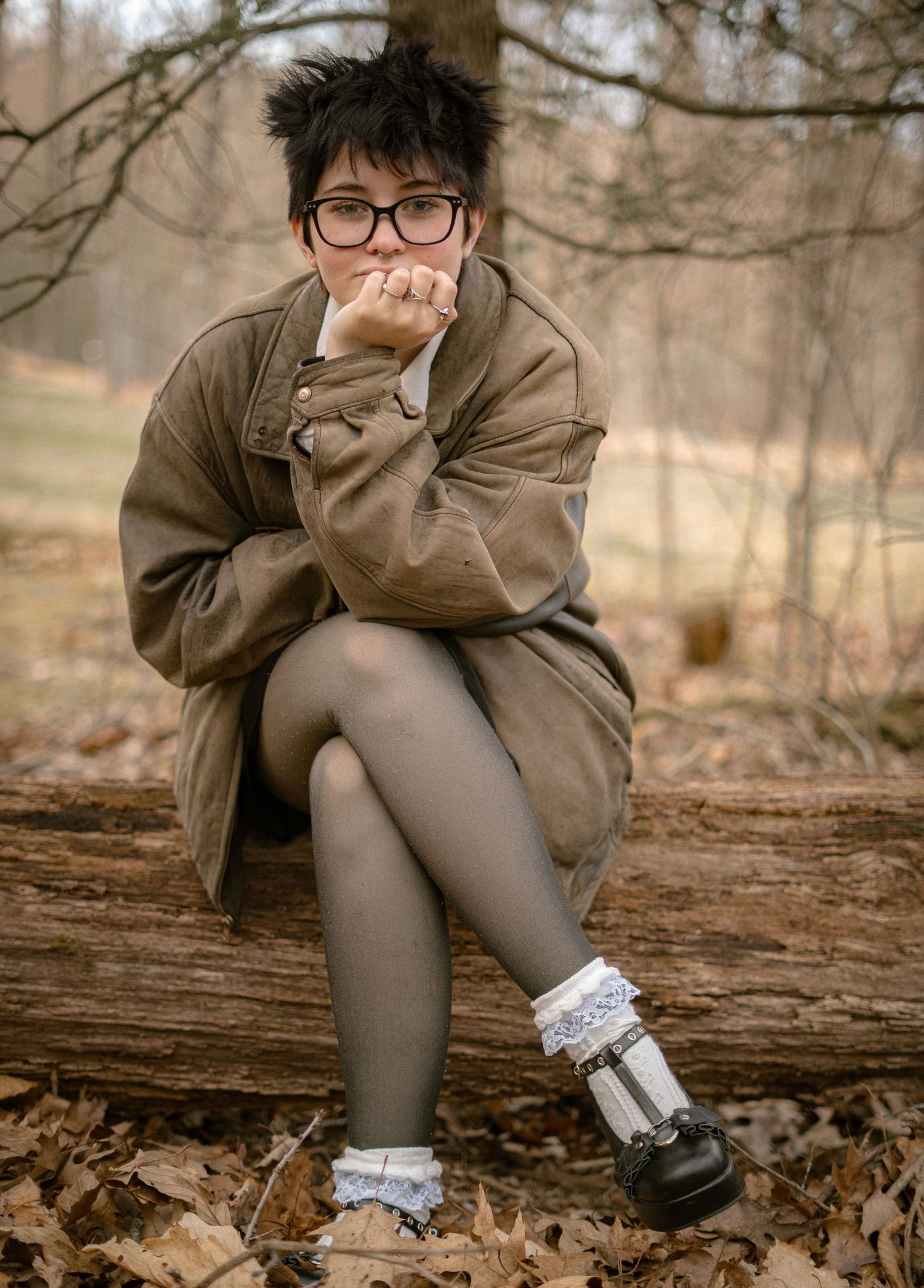 Someone wearing a brown coat and glasses sits thoughtfully on a fallen log in an autumn forest setting.